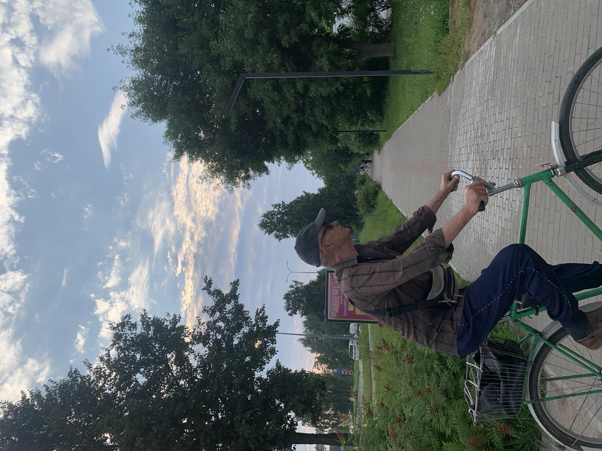An elderly man wearing a brown plaid shirt, dark pants, a grey cap, and glasses is riding a green bicycle on a paved bike path. He is in profile, looking to the right. The bicycle has a wire basket on the back containing black items. A man's shoulder bag is slung across his chest. The bike path is lined with green grass and trees on both sides. In the background, there are more trees, streetlights, and a billboard with Russian text. Cars are visible on a road to the left of the bike path. The sky is blue with scattered clouds. The scene appears to be in the city of Chaikovsky, Russia.