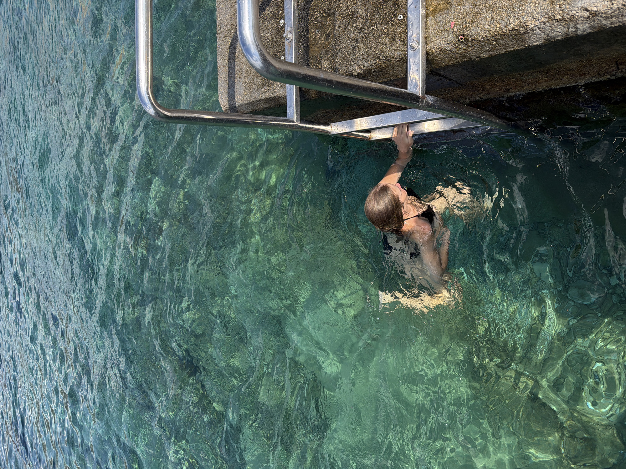 A person is partially submerged in clear, turquoise water next to a concrete structure with a metal ladder. The person, viewed from above, is wearing a black swimsuit and has their right hand resting on the ladder's railing. Their head is turned away from the camera, and their hair is wet, suggesting they have just been swimming or are about to enter the water.
The water is calm, with sunlight reflecting off its surface, creating dappled patterns on the bottom. The concrete structure appears to be a pier or a seawall, with visible rivets or bolts securing the ladder. The scene suggests a warm, sunny day, likely in a coastal or lake setting. The clear water indicates good visibility and possibly a shallow depth near the edge. The overall mood is peaceful and serene, inviting a sense of leisure and enjoyment of the water.
The location is identified as Budva, Montenegro. There is no visible text in the image.