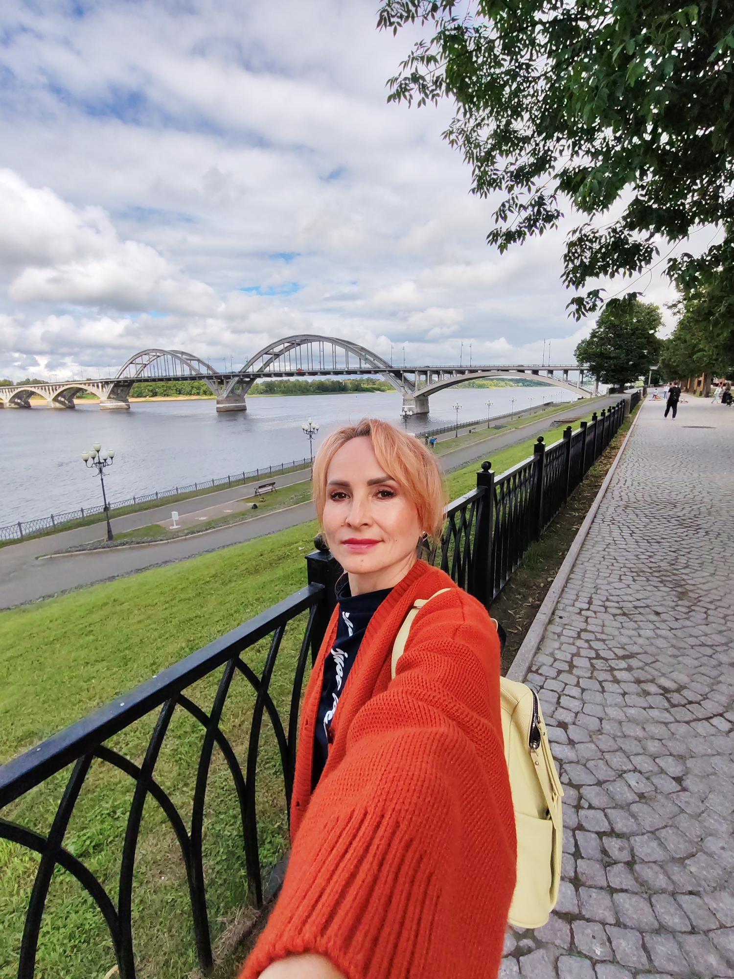 A woman with short, reddish-blonde hair and red lipstick is taking a selfie with a bridge and a river in the background. She is wearing an orange cardigan over a dark top with white lettering and a light yellow backpack. The woman is standing on a cobblestone walkway next to a black metal fence. To her left, a grassy slope leads down to a riverbank with lampposts and a path. In the background, a multi-arched bridge spans the river. The sky is cloudy. The image appears to be taken in the city of Bol'shie Mkhi, Russia, as indicated by the presence of the distinctive bridge.