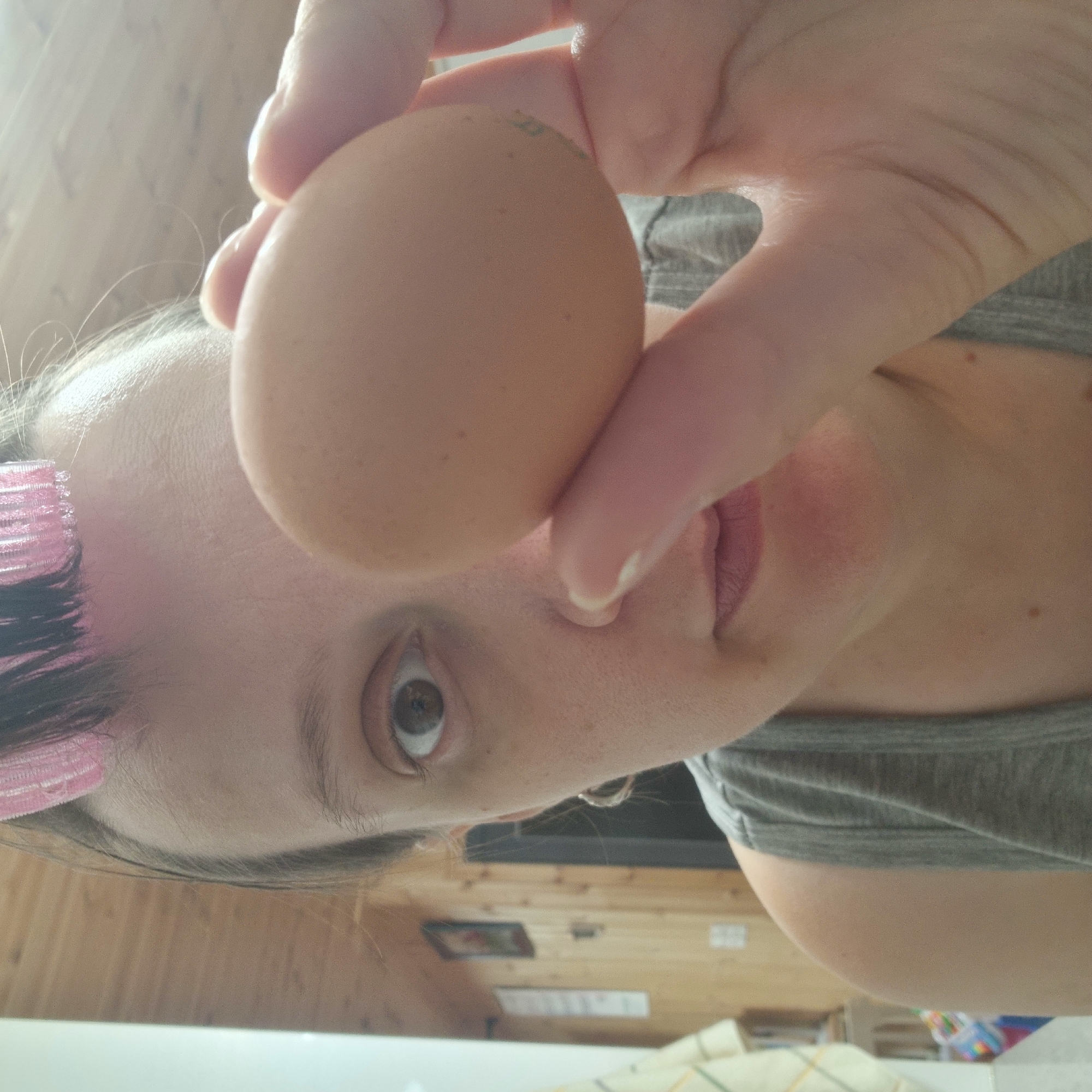 The image is a close-up, first-person perspective photograph of a person holding a light brown egg in front of their face. The person is wearing a grey tank top and has pink hair rollers in their hair. Their left eye is visible and appears to be looking at the camera. The background is a wooden paneled wall with some framed pictures and a light-colored cloth hanging on the left. The overall lighting is bright, suggesting it might be daytime. The image is consistent with a casual, at-home setting, possibly in a kitchen or bedroom. There are no visible landmarks or specific features that would indicate a precise location within Dunnae-myeon, South Korea.
