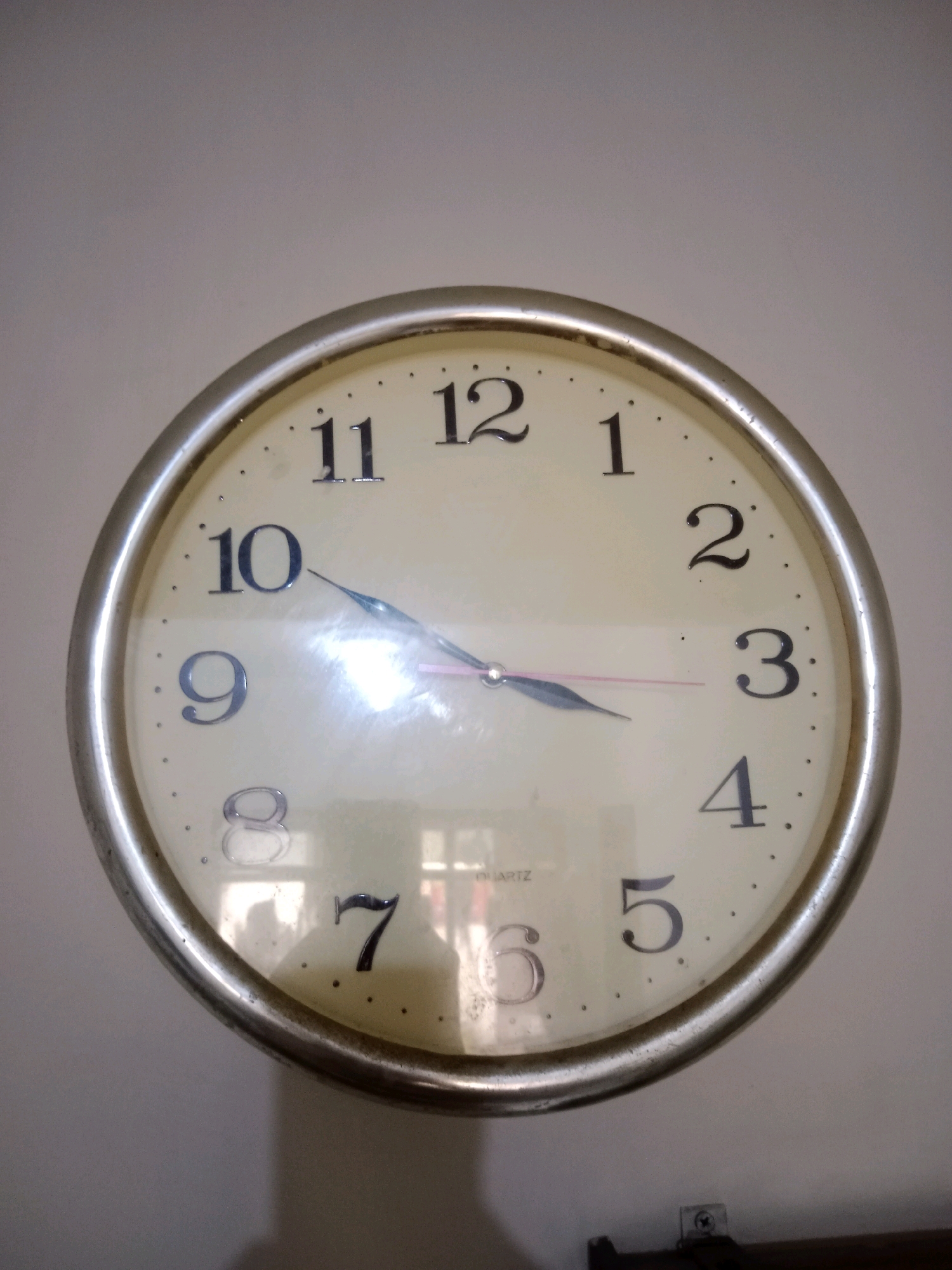 A close-up, eye-level, medium shot captures a round analog clock mounted on a light-colored wall. The clock has a metallic silver rim and a cream-colored face with black Arabic numerals indicating the hours. The clock hands, one silver and one pinkish-red, point to approximately 9:47. The word "QUARTZ" is visible near the 6 o'clock position. The lighting suggests the image was taken indoors, possibly during the day, with some glare reflecting off the clock face. A faint shadow of a person is visible on the lower right quadrant of the clock face. Below the clock, a dark horizontal object, possibly a shelf or fixture, is partially visible. The background is a plain, textured wall. The image appears to be taken in Batu, Indonesia.