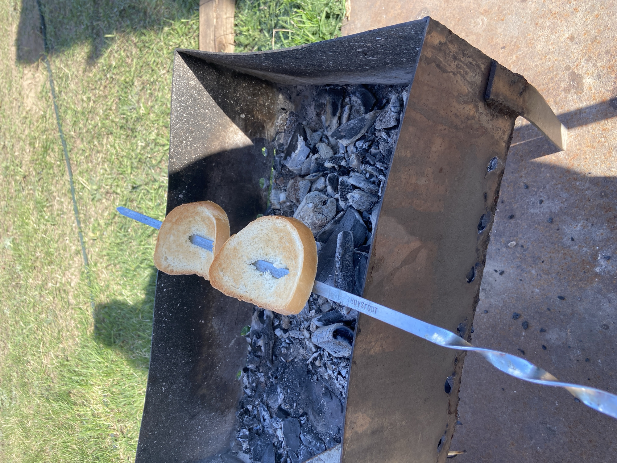 A metal skewer with two slices of bread impaled on it is positioned over a barbecue grill containing charcoal embers. The grill is located outdoors on a surface with grass and a concrete or stone-like area. The word "BOYSCOUT" is visible on the skewer. This location is in the city of Ольховка, Russia.
