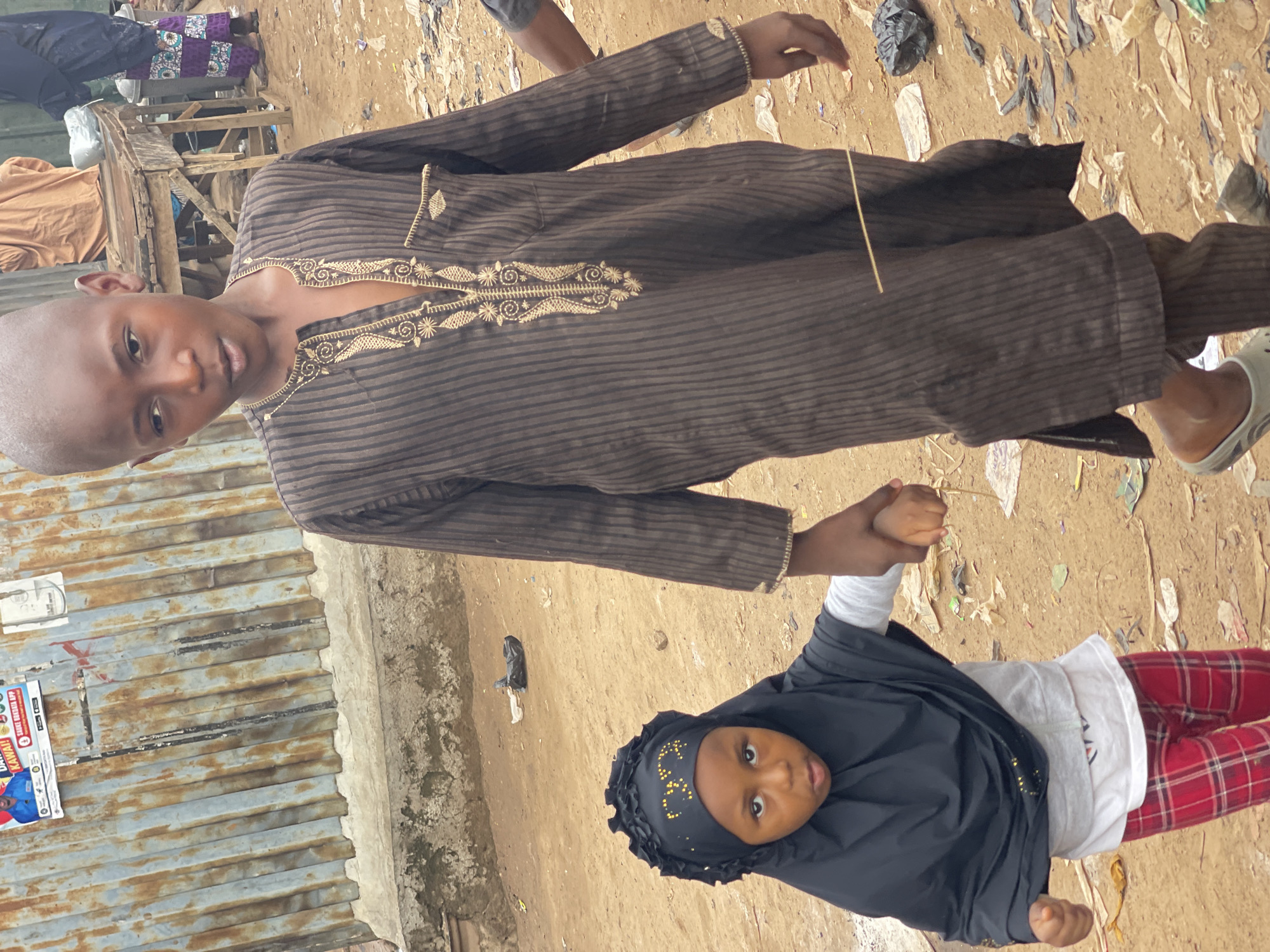 In Chikaji, Nigeria, a young boy in a striped brown robe and sandals holds the hand of a young girl wearing a black hijab, white shirt, and red plaid pants. They stand on a dirt ground covered in trash. To the left is a corrugated metal wall with a poster. In the background is a wooden structure with various items on top.