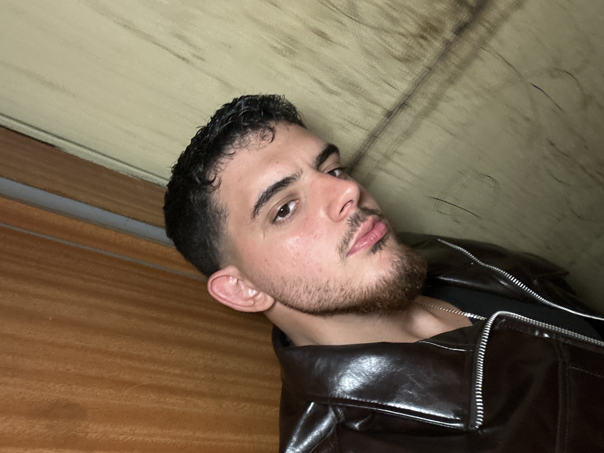 A medium shot depicts a man with dark, wavy hair and a beard. He is wearing a dark brown leather jacket with silver zippers and is positioned at an angle, looking towards the viewer. The background consists of a light beige, possibly wooden, surface. The image does not provide enough information to determine the location in Lovech, Bulgaria.
