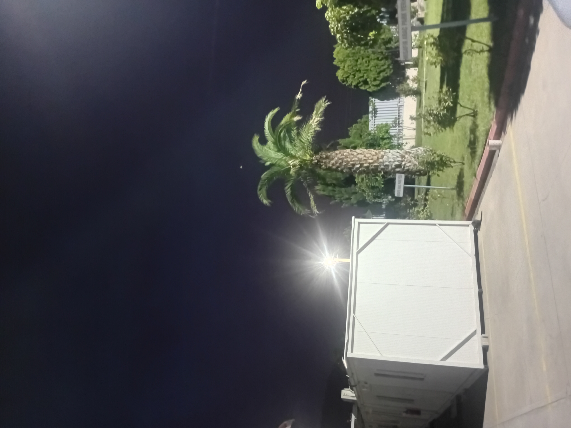 A mobile building, possibly a temporary office or dwelling, is situated on a paved lot at night. The building is white with visible paneling and is elevated slightly off the ground by concrete blocks. To the right of the building, a palm tree stands tall amidst other greenery. Several signs with Turkish text are visible in the landscaped area to the right, suggesting this location might be in the city of Onikişubat, Turkey. A bright light source, possibly a floodlight, illuminates the scene, casting a strong glare. The sky above is dark, indicating it is nighttime. There are no people visible in the image.