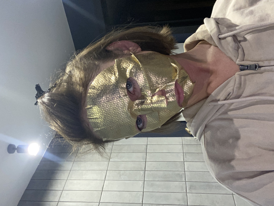 An individual is depicted in a close-up, chest-up indoor photograph taken in Ust-Ilimsk, Russia. The subject is wearing a gold-colored sheet face mask with a textured, honeycomb-like pattern covering the majority of their face, including the forehead, nose, cheeks, and chin. Blue eyes are visible through the mask's eye openings, and the mouth area is also exposed. The subject has short to medium-length light brown hair, secured partially at the crown with a black claw clip. They are wearing a light beige or cream-colored hooded sweatshirt (hoodie) with a silver zipper down the front, which is partially unzipped. The background consists of a light gray tiled wall with dark vertical grout lines, with a darker, untextured surface visible above the tiles. A black cylindrical light fixture emitting bright light is mounted on the upper wall or ceiling in the upper left quadrant of the frame.