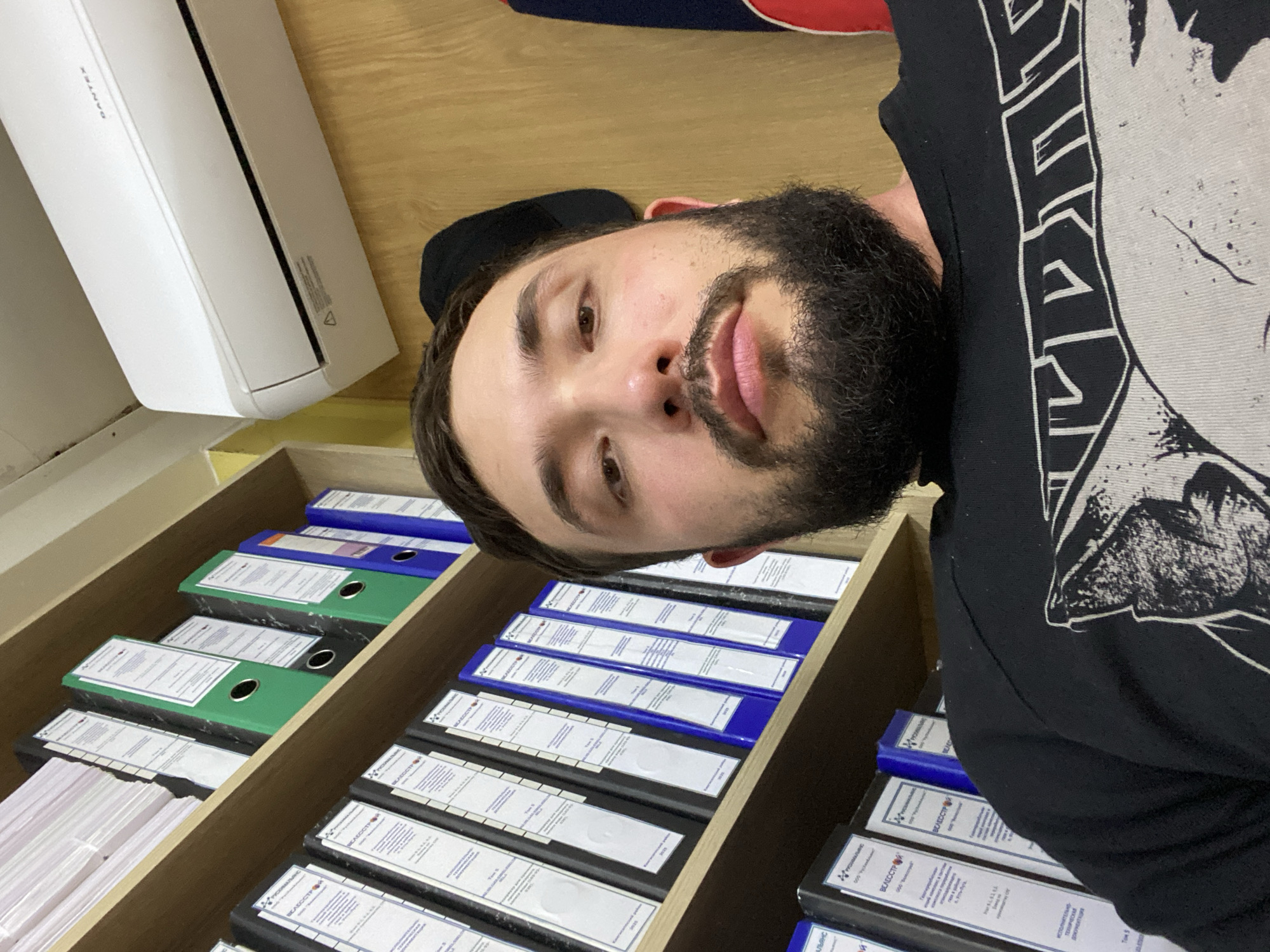 The image shows a close-up selfie of a man with a beard and dark hair, wearing a black t-shirt with a graphic print. He is indoors, with shelves filled with binders and files visible behind him. An air conditioning unit is mounted on the wall above the shelves. The background suggests an office or storage environment. The scene is set in Luzhitsy, Russia.