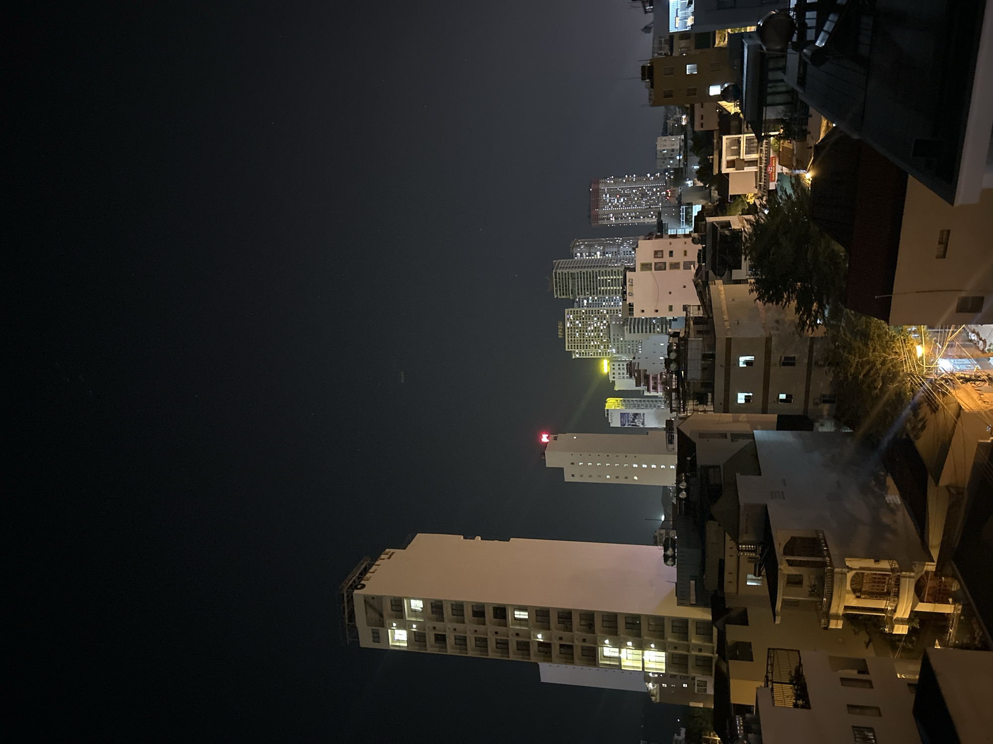 Night cityscape in Bắc Nha Trang, Vietnam. High-rise buildings with illuminated windows dominate the skyline, while smaller buildings and residential structures fill the foreground. The sky is dark, with scattered lights from distant buildings. A few illuminated streets can be seen.
