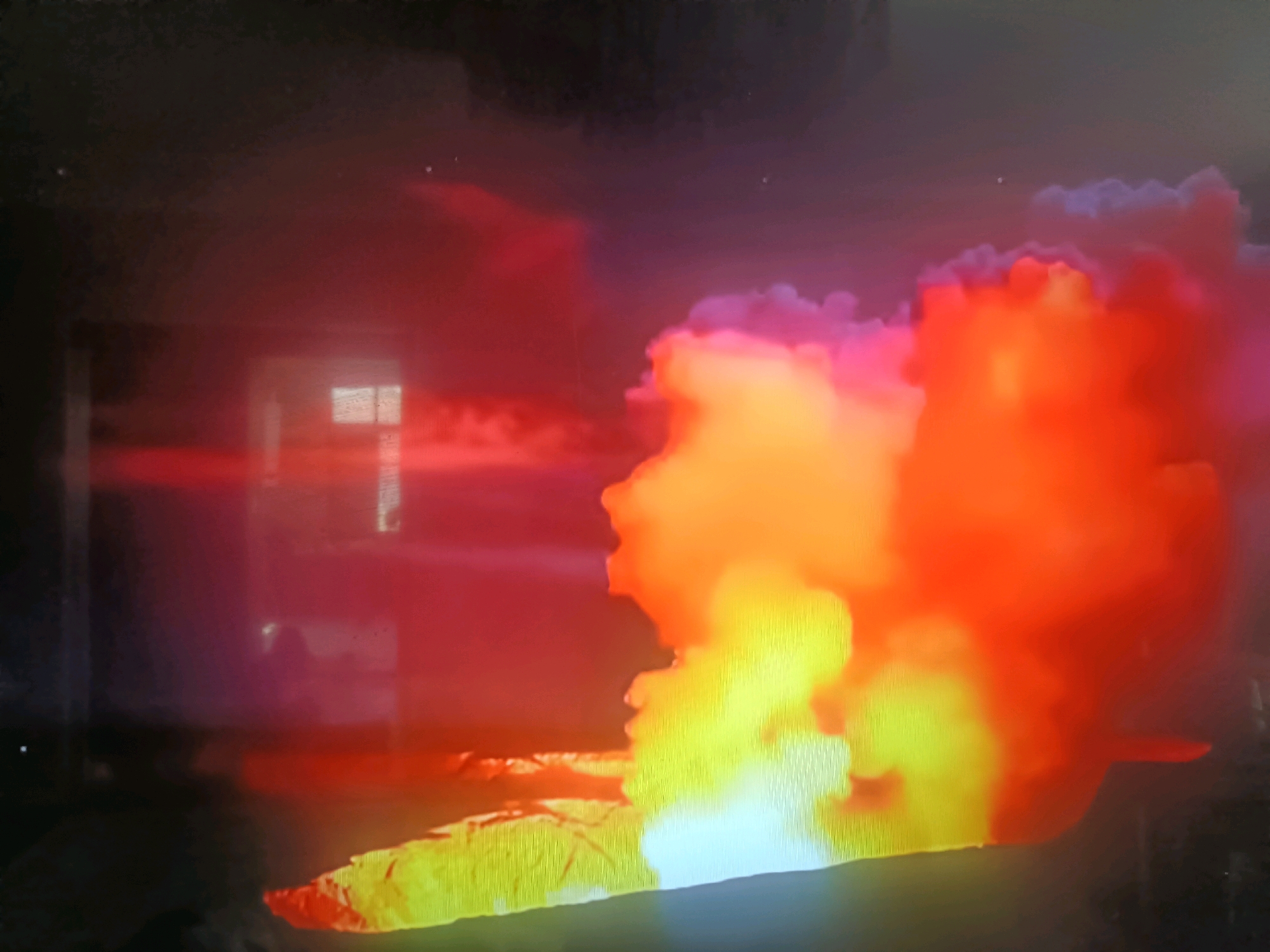 The image shows a chaotic scene of intense heat and light, likely an industrial accident or a fire. A large plume of bright yellow and orange smoke billows upwards and to the right, obscuring any background detail except for a dark, presumably overcast sky with faint celestial bodies. To the left, a building with illuminated windows is partially visible, suggesting human presence or proximity to a populated area. The overall impression is one of destruction and uncontrolled energy, possibly a large-scale fire or explosion. The scene does not provide any specific clues to pinpoint its location in Қасымбек датқа, Kazakhstan, nor does it depict any individuals or specific actions.