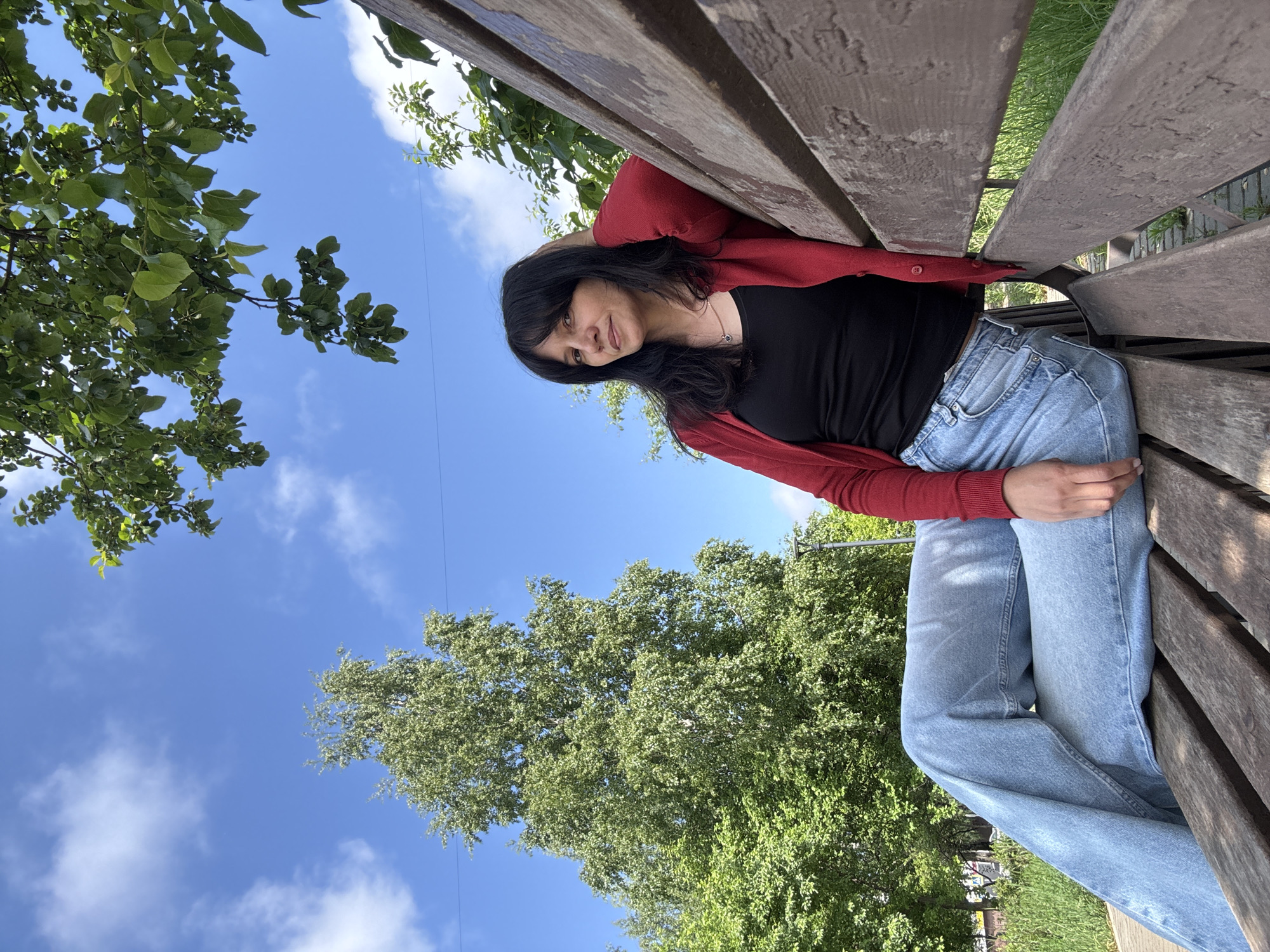 A young woman with dark hair is seated on a wooden park bench, captured outdoors on a bright, sunny day. She is wearing a red long-sleeved cardigan over a black top and light blue jeans. Her posture is relaxed, with her head slightly tilted and a gentle, soft smile on her face as she looks forward. Her right arm rests on her leg, while her left arm appears to be behind her head or along the back of the bench.
The setting features a clear blue sky adorned with scattered white clouds, indicating pleasant weather. Lush green trees and foliage are prominent in the background, suggesting a park or a natural outdoor area. The wooden bench shows signs of age and wear. The overall impression is one of calm and tranquility. No visible text is present in the image.