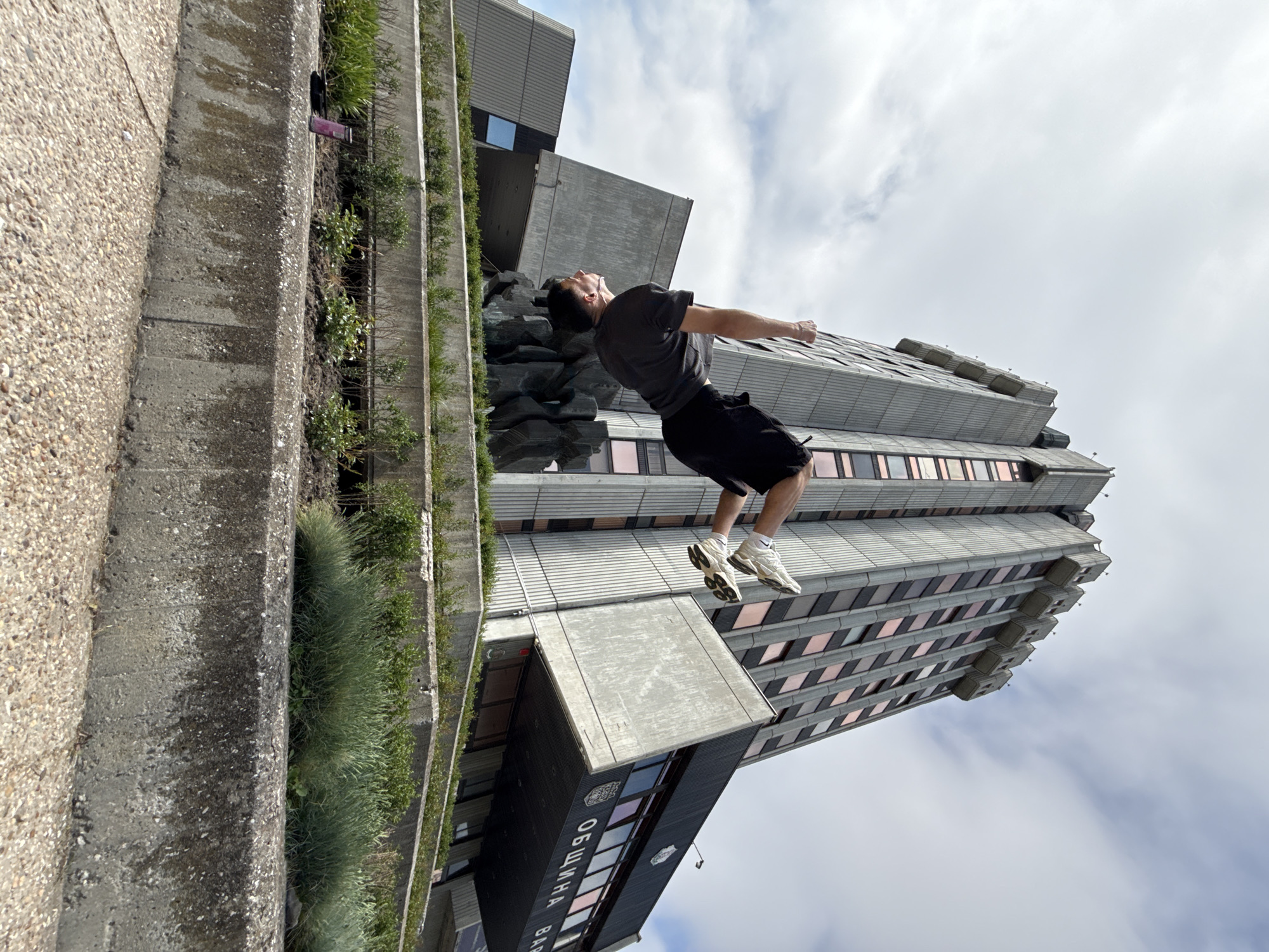 A man in mid-air performing a backflip stands out in front of a high-rise building with multiple levels and many windows. The man is wearing a gray shirt, dark shorts, and white sneakers. He is angled back, with his arms outstretched and head looking up. The building behind him has a gray facade with horizontal lines and is topped by stepped sections. To the left, there is a concrete structure with tiered planters filled with greenery. The sky is overcast. The building is identified by text as "Община Варна", which translates to "Municipality of Varna," placing the scene in Varna, Bulgaria.