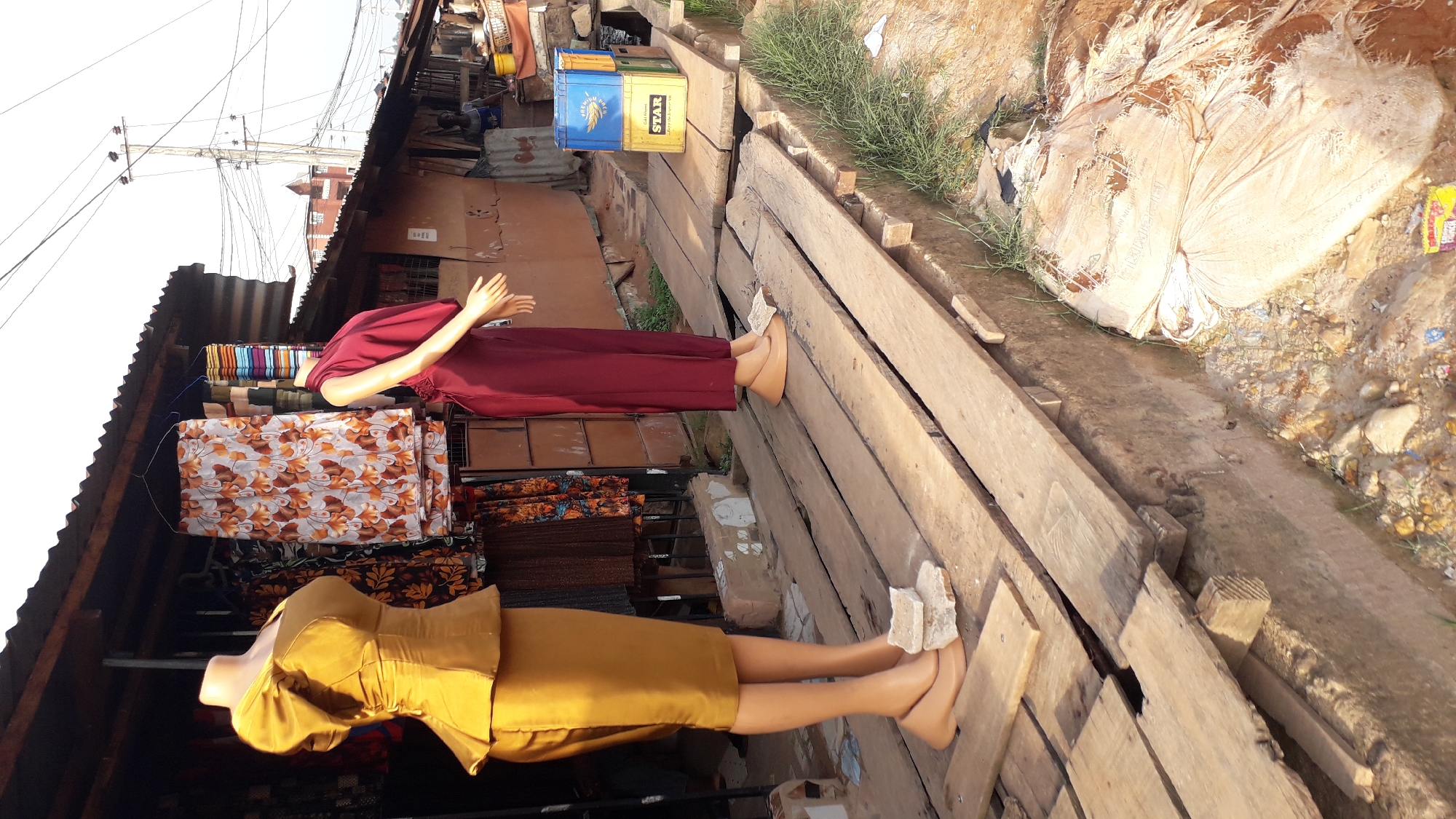 The image depicts a street scene in Nkpor, Nigeria, featuring two mannequins displayed outside a fabric shop. The mannequins are positioned horizontally on a makeshift wooden platform, each adorned with different garments. The lower mannequin wears a golden yellow dress, while the upper mannequin is dressed in a maroon colored outfit.
The shop's interior showcases rolls of fabrics in various colors and patterns. Adjacent to the wooden platform is a drainage ditch with trash. The surroundings include buildings and overhead electrical wires.