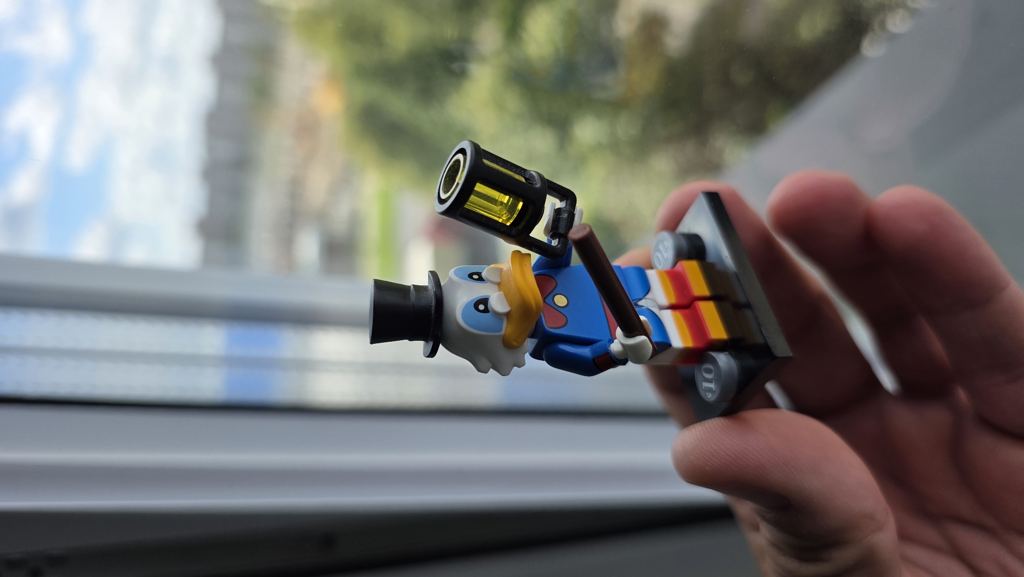 A Lego minifigure of Scrooge McDuck, wearing a top hat, blue coat, bow tie, and striped pants, is held in a person's hand. The minifigure holds a brown cane with a yellow lantern attachment. The background shows a blurred view of buildings and trees through a window with white curtains. There is no information about the city of Brest, Belarus, in the image.