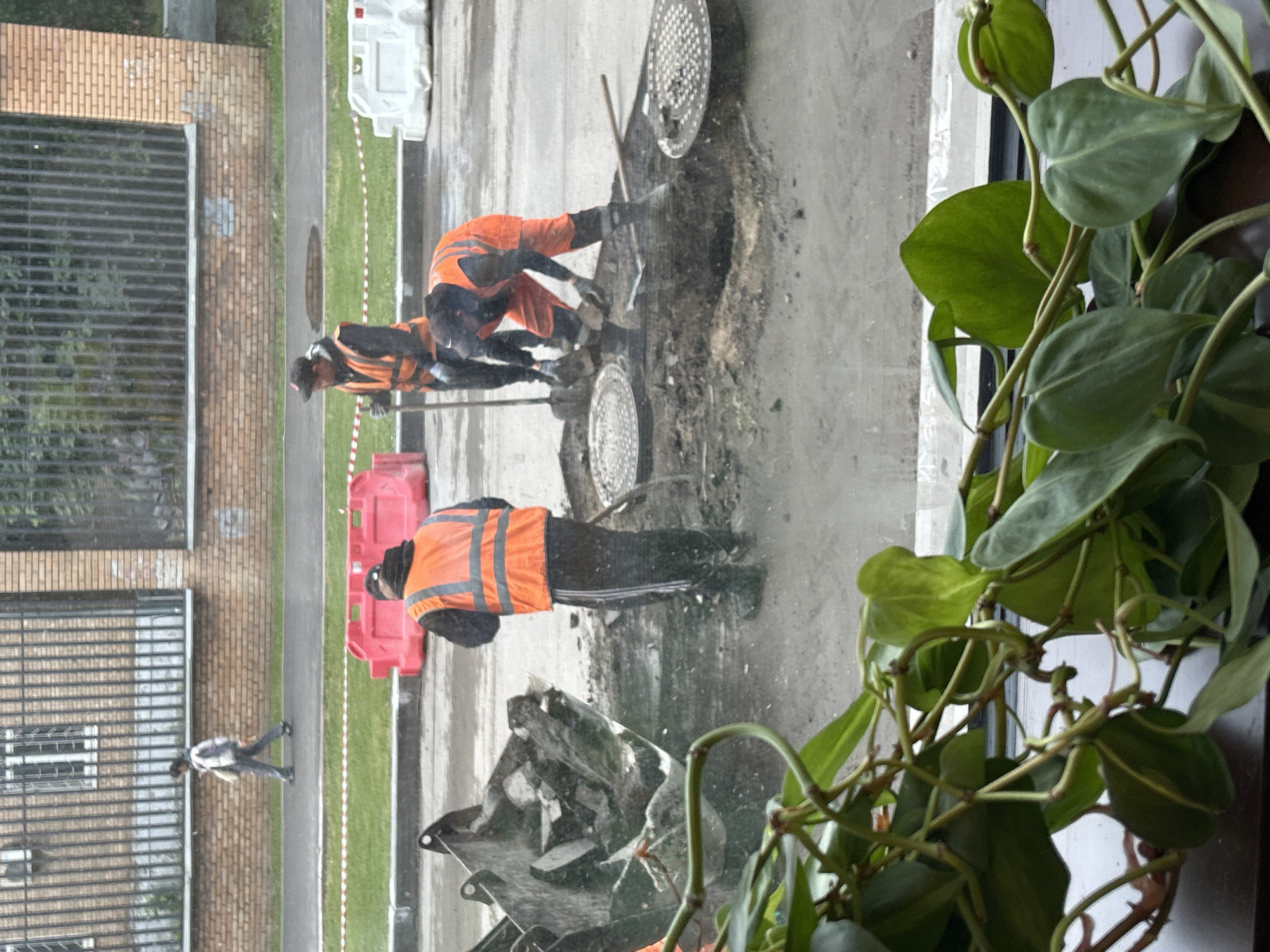The image depicts a scene outside a building in an urban environment, as viewed through a window with a potted plant in the foreground. In the street, there are two workers wearing orange safety vests working near an open manhole. A third worker is standing nearby. Nearby are red plastic barriers. The street surface appears to be wet or damp. A pedestrian is walking away from the scene. The building visible through the window has a brick facade with barred windows.