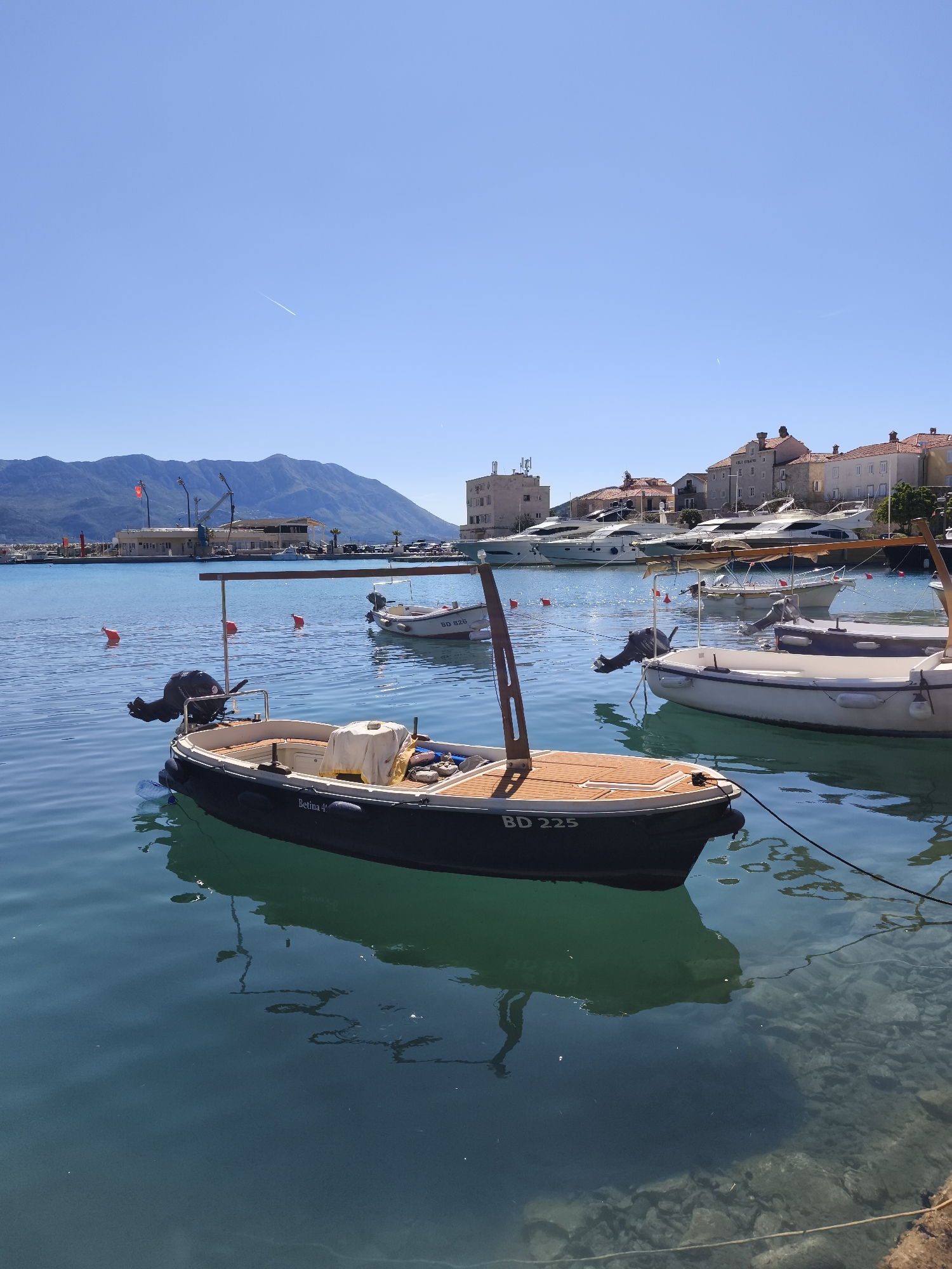 The image depicts a marina in Budva, Montenegro on a clear day with a cloudless blue sky. In the foreground, a small boat, labeled "Betina 4" and "BD 225", is moored. This boat features a dark hull, wooden decking, and a rudimentary canopy frame. To its right are other similar boats, also moored. In the background, across the tranquil water, are buildings of Budva's old town, characterized by their reddish roofs and light-colored facades. A mountain range is faintly visible further in the background. Red mooring buoys are scattered in the water. The water is clear enough to see the bottom in some areas. The overall atmosphere is calm and sunny.