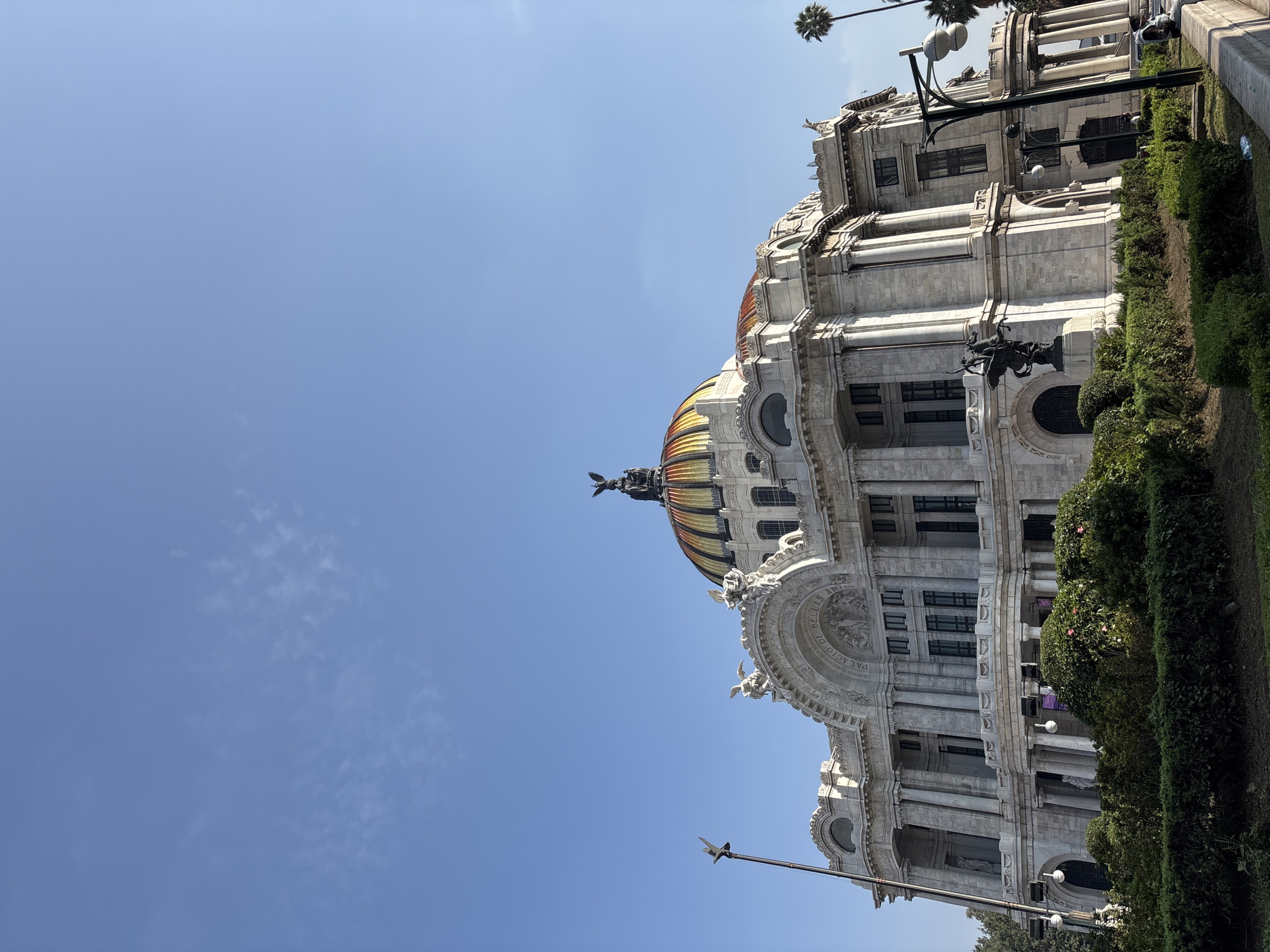 This is a low-angle, outdoor, daytime, eye-level, full-body shot of the Palacio de Bellas Artes in Mexico City, Mexico. The building is made of marble and features a dome with gold and red accents. A statue of a winged figure stands atop the dome, and other statues adorn the building's facade. Lush green bushes and hedges are planted in front of the building, and palm trees are visible in the background. The sky is a clear blue with a few wispy clouds. A lamppost with two lights stands on the right side of the frame.