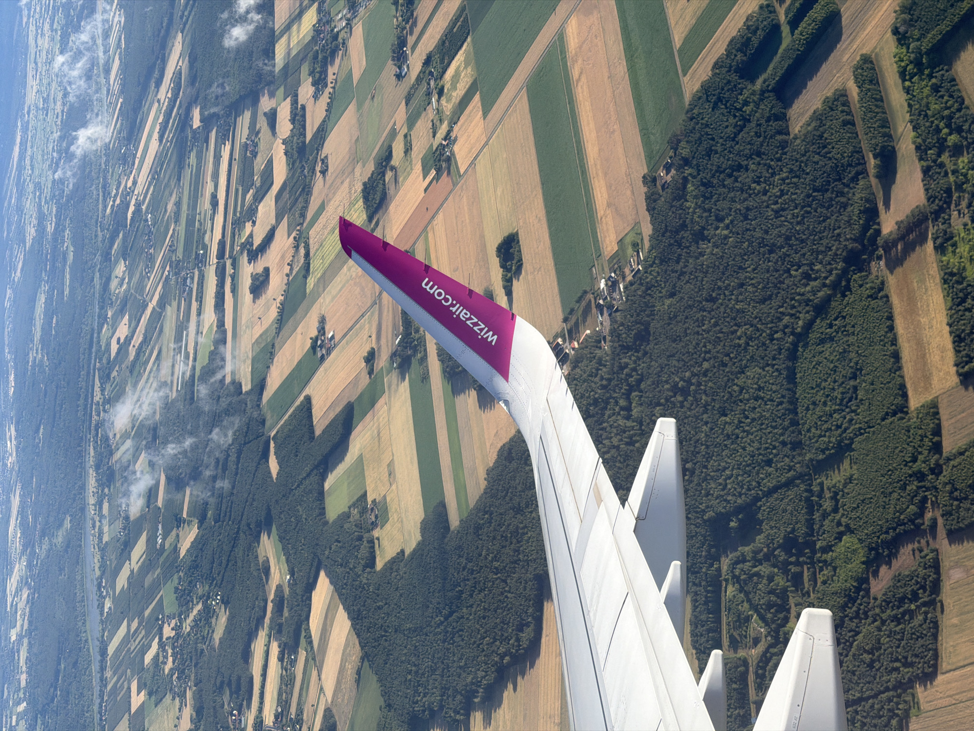 An aerial, slightly oblique view captures an airplane wing as it flies over a rural landscape. The wing, predominantly white with a prominent pink accent featuring the "wizzair.com" logo, dominates the foreground. Below, a patchwork of agricultural fields displays varying colors of green, yellow, and brown, interspersed with dark green forest areas. Scattered houses and roads are visible within the fields and along the edges of the forests, indicating a sparsely populated rural area. The scene suggests a clear day with some wisps of clouds visible in the distance. The location is identified as Ziąbki, Poland.