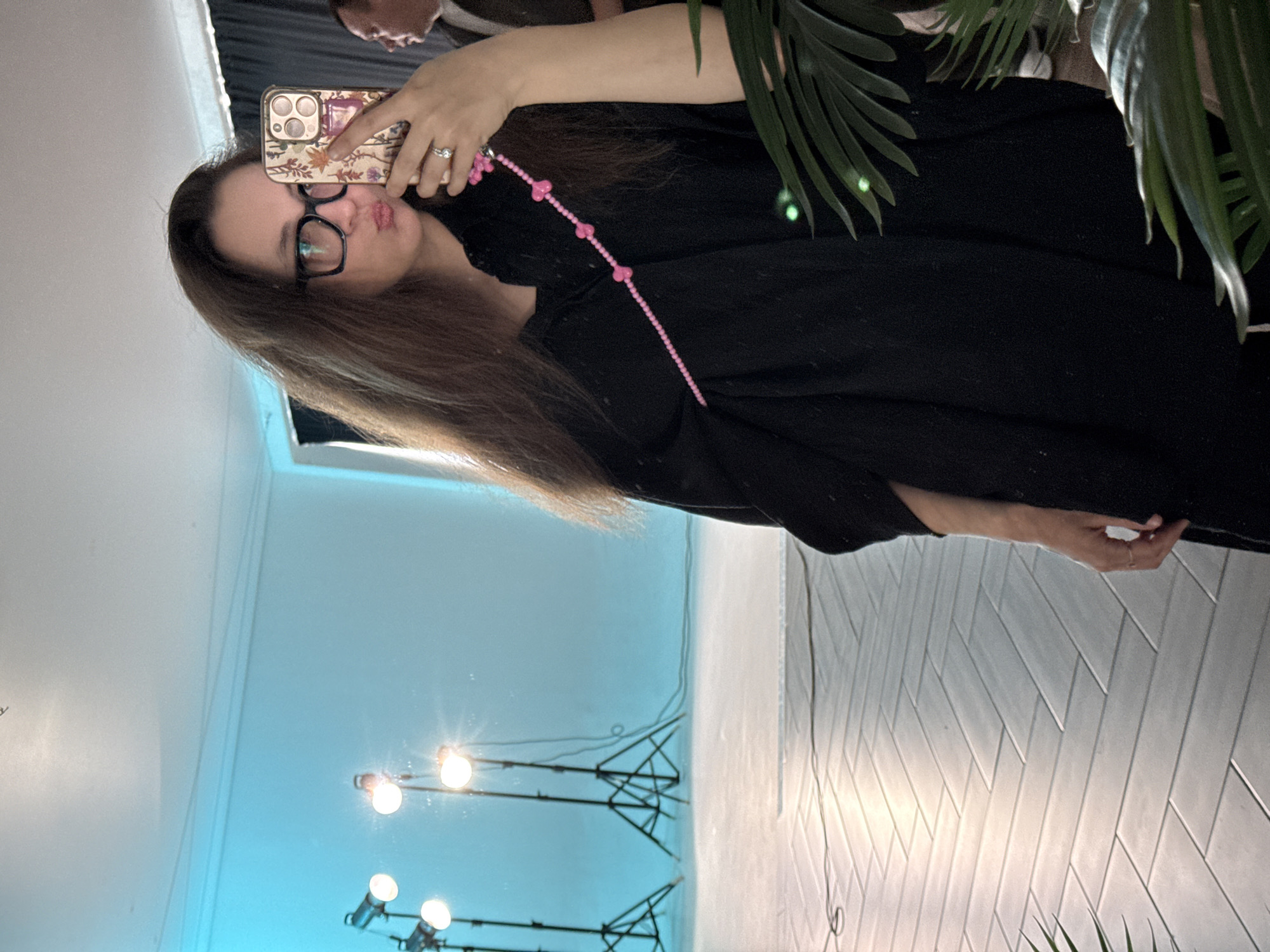A woman with long brown hair and glasses holds a smartphone and takes a selfie in front of a mirror. The mirror reflects a room with a turquoise wall, studio lights, and a light-colored floor with a herringbone pattern. The location is unknown and there is no mention of Russia or Chaikovsky.