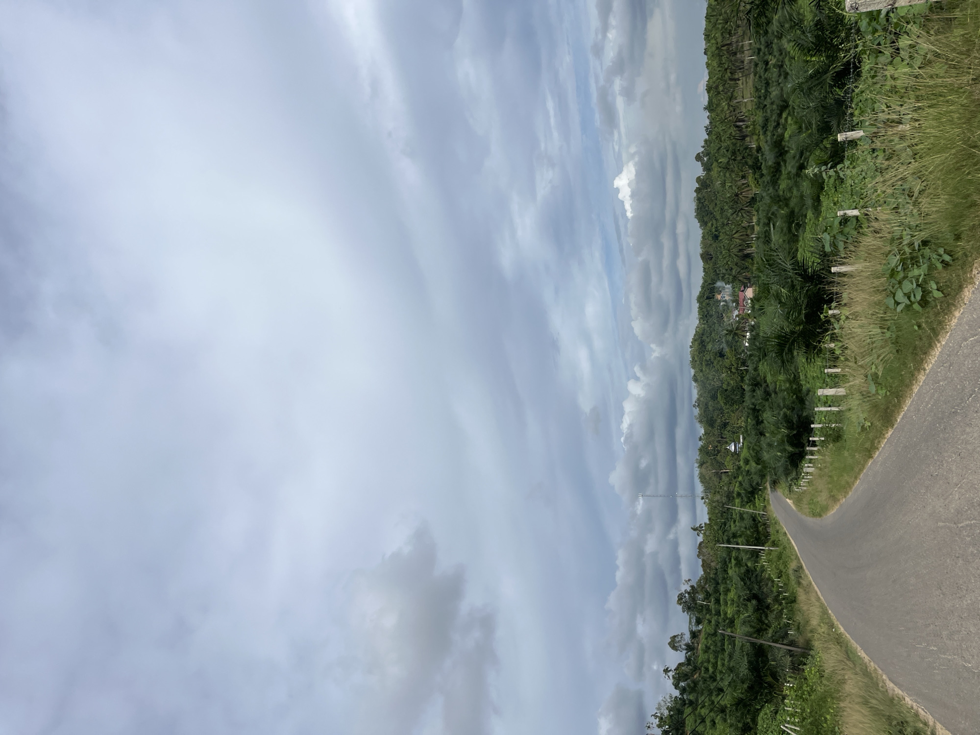 The image depicts a rural road winding through lush green vegetation, likely palm trees, under a cloudy sky. In the distance, a tall communication tower and a white dome-shaped structure, possibly a mosque, are visible. The road is paved and appears to be in good condition, with a dry grass verge and simple wooden post fencing on either side. The scene conveys a sense of tranquility and isolation, characteristic of a remote area. The location is identified as Sukajadi, Indonesia.