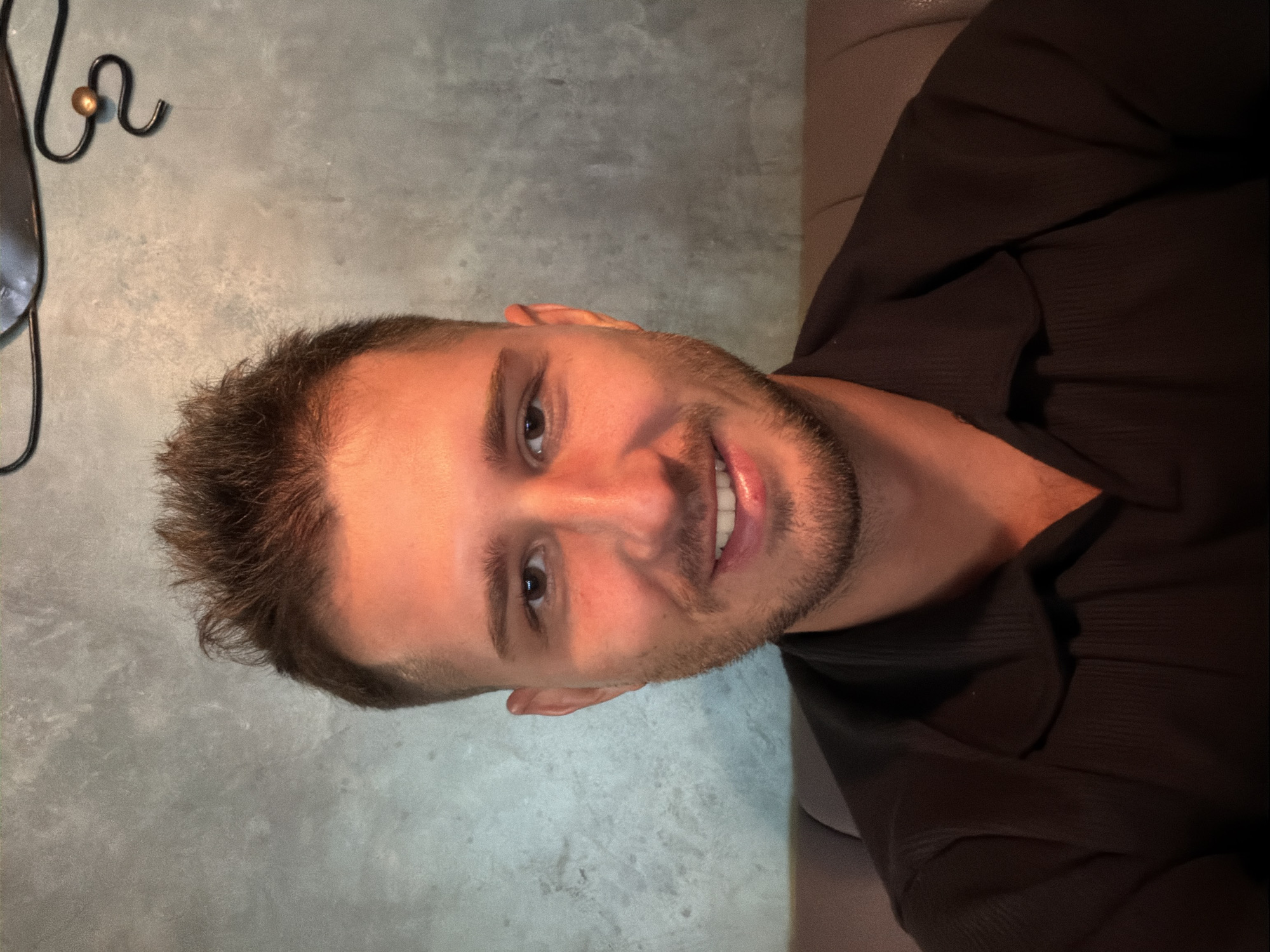 A man is looking at the camera. He is wearing a dark brown shirt and has short brown hair. The background is a grey wall. The location is not discernible from the image, and Moscow, Russia cannot be determined from the provided image.