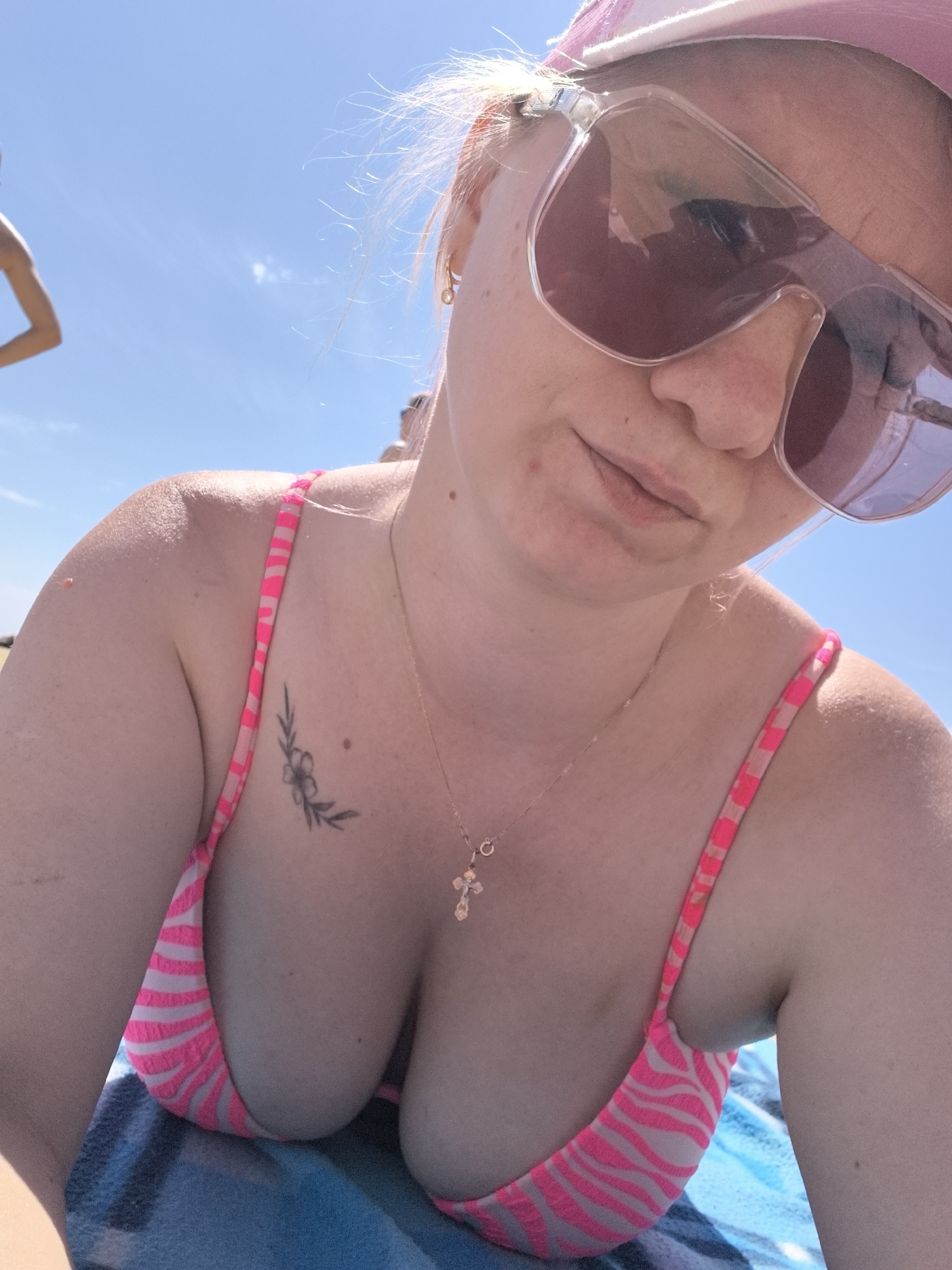 A close-up shot captures a woman in a pink and white striped bikini top, resting on a blue towel under a clear blue sky, suggesting a beach location in Anapa, Russia. She wears large pink-tinted sunglasses and a pink baseball cap. A delicate gold necklace with a cross pendant is visible around her neck, resting on her cleavage. A small floral tattoo adorns her left shoulder. Her hair, light blonde, is slightly windswept. The background reveals a hint of another person walking in the distance on the left, also against the bright sky. The lighting suggests a sunny day.