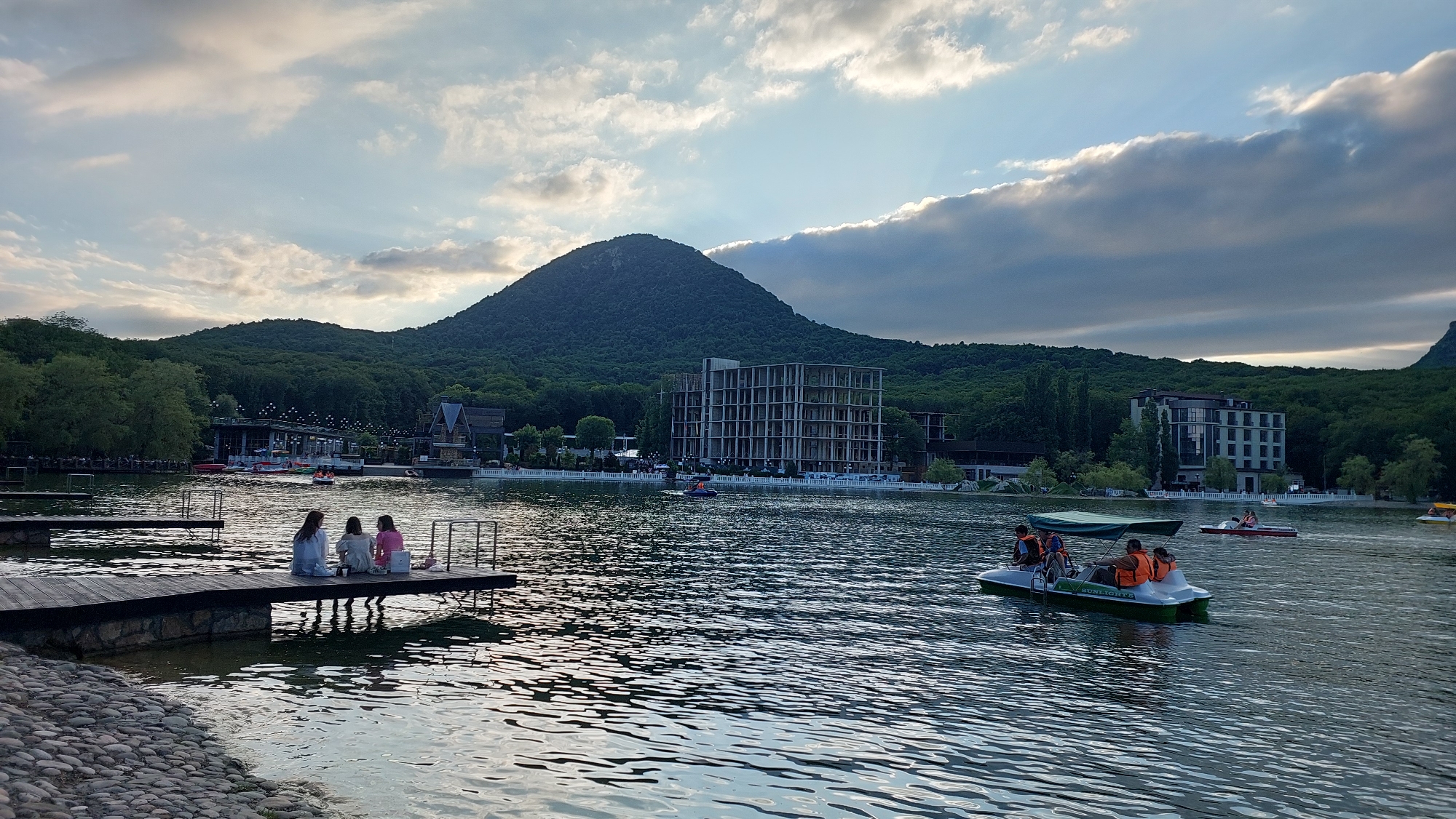 This media file captures a serene lake scene in Zheleznovodsk, Russia, during what appears to be late afternoon or early evening. The sky is dramatic with a mix of illuminated clouds and darker, overcast formations, suggesting dynamic weather. A large, tree-covered mountain dominates the background, casting a shadow over a resort-like area with modern buildings and traditional architecture lining the lake's edge.
On the lake, several pedal boats are in motion, with people enjoying a leisurely ride. One pedal boat in the foreground has four occupants, all wearing orange life vests, and the boat itself has "SUNLIGHTS" printed on its side. Another pedal boat further back also has people wearing life vests. In the middle ground, a few smaller boats are visible, and people can be seen along the shore and on piers.
In the foreground, a wooden pier juts out into the water, with three young women seated on it, facing the lake. They appear relaxed, and one is looking at a phone. The pier is built on a stone foundation, and the edge of the lake is lined with rounded stones. The overall mood is peaceful and recreational, highlighting a popular tourist destination.