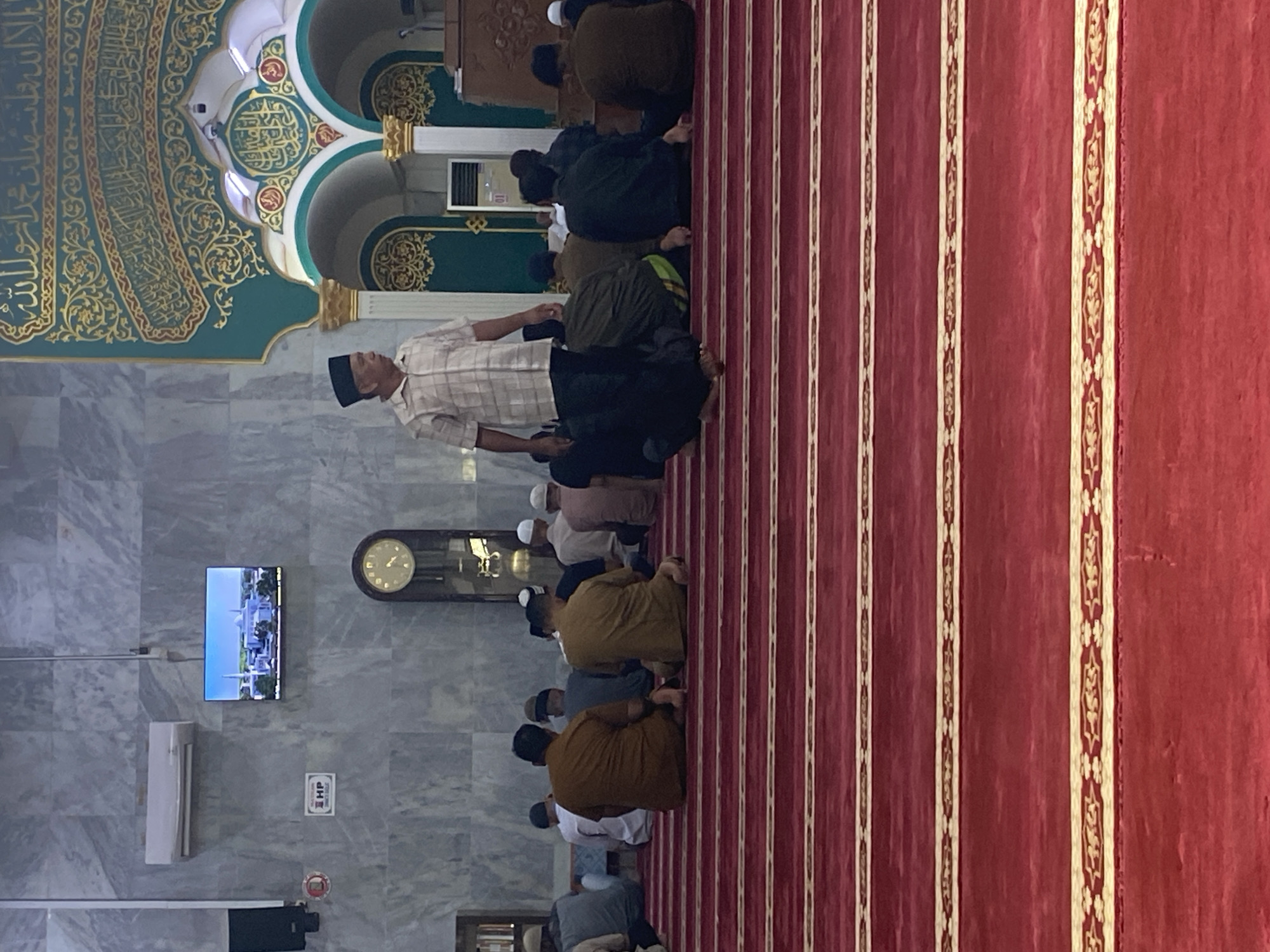 A man in a checkered shirt and a black cap stands in the middle of a mosque, facing to the right. He is in the foreground of the image, which shows a group of men sitting on a red carpet in prayer. The mosque has marble walls, and a large clock hangs on the wall behind the praying men. A television screen on the wall shows an image of a mosque. The scene is illuminated by artificial light. The image was taken in Lhokseumawe, Indonesia, specifically at the Masjid Raya Baiturrahman.