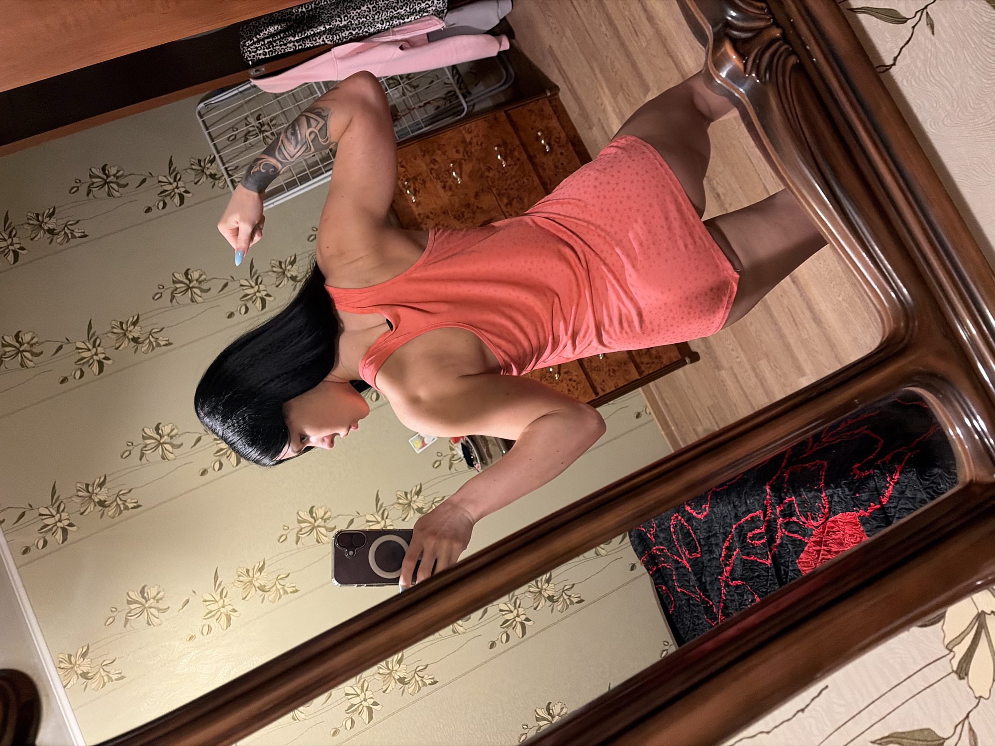 A young woman with black hair is taking a mirror selfie in a room. She is wearing an orange tank top and flexing her bicep. She has a tattoo on her left arm. The room has floral wallpaper and a wooden dresser. In the reflection, a black blanket with red floral patterns is visible on a bed. The scene is indoors and appears to be in a residential setting. No information about the city of Ufa, Russia is visible or inferable from the image.