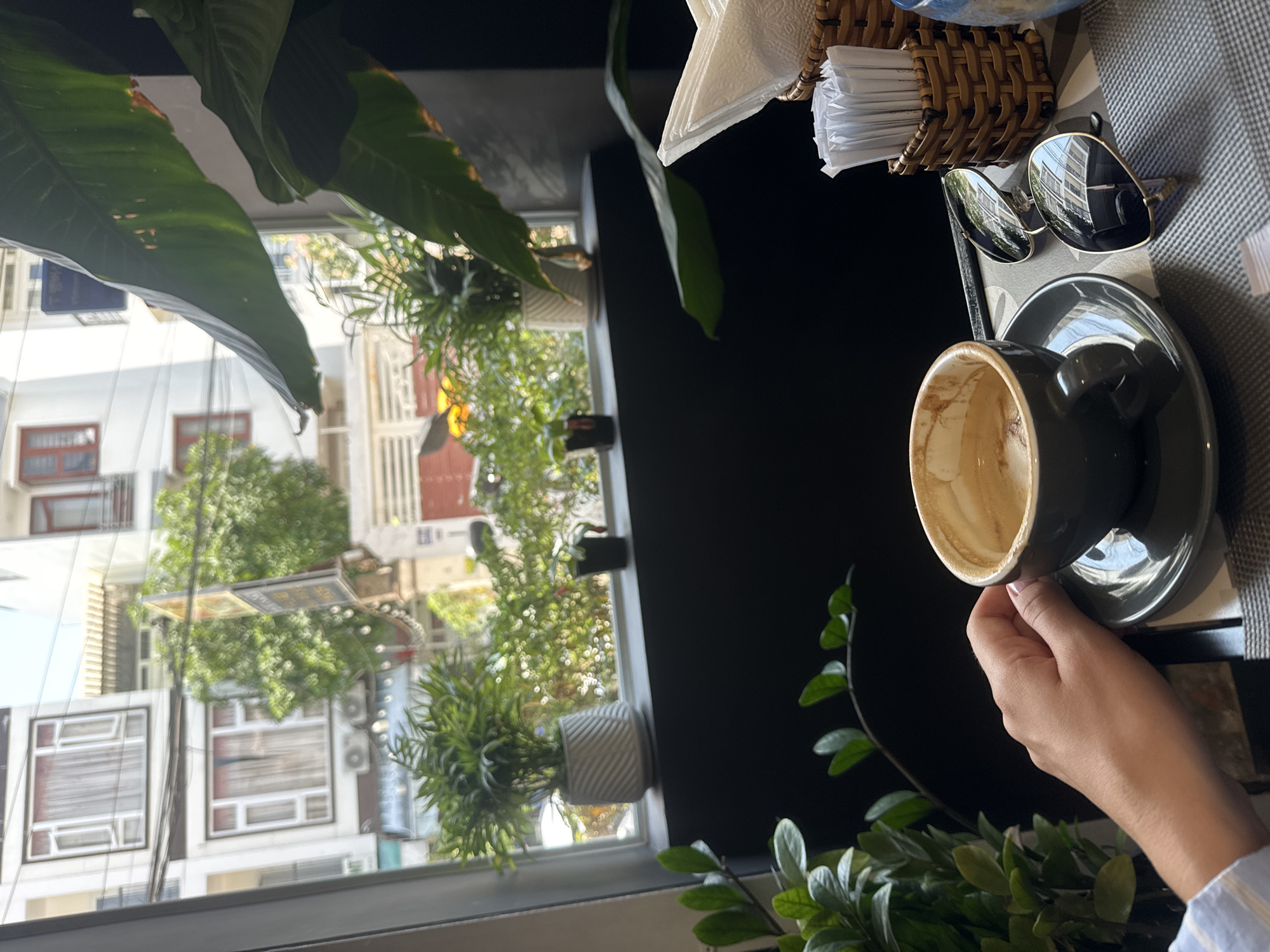 A partially-consumed coffee cup rests on a saucer, held in a person's hand. Sunglasses and a napkin holder sit nearby on a table. A window in the background shows an urban street scene with multi-story buildings and greenery. Plants are situated near the window, both inside and outside. The location appears to be a cafe in a city.