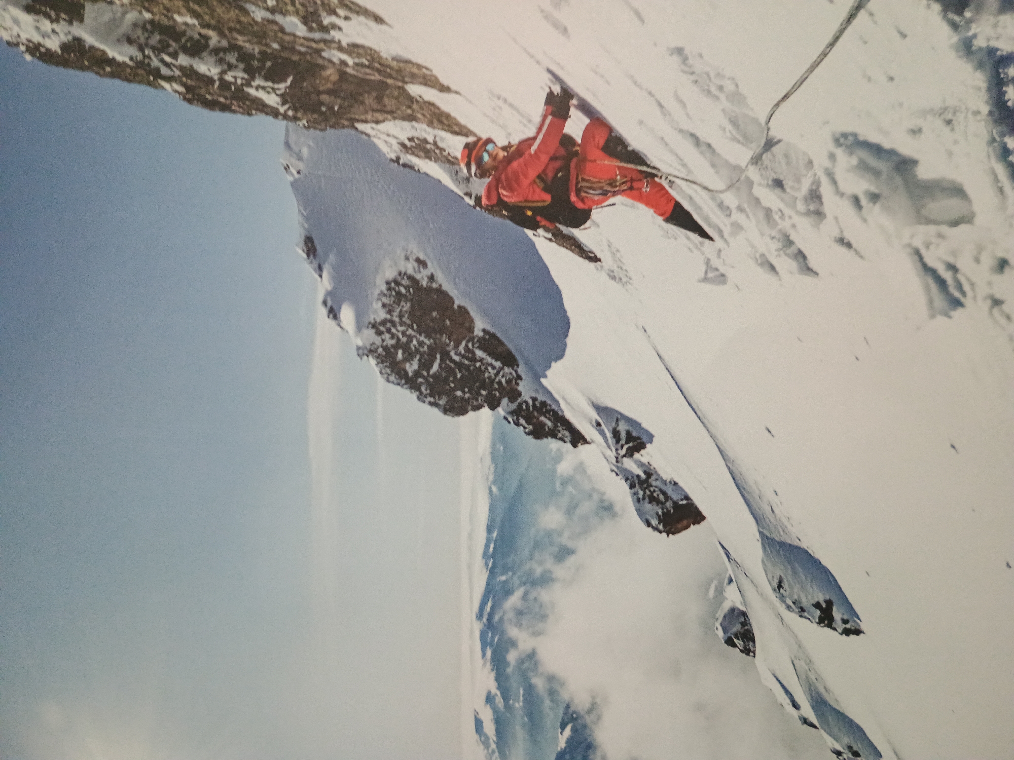 A mountaineer in red climbing gear is traversing a steep, snow-covered mountain face. The scene includes rock formations, a rope, and a sky partly obscured by clouds. The location is mountainous terrain, likely a high-altitude environment. There is no identifiable city.