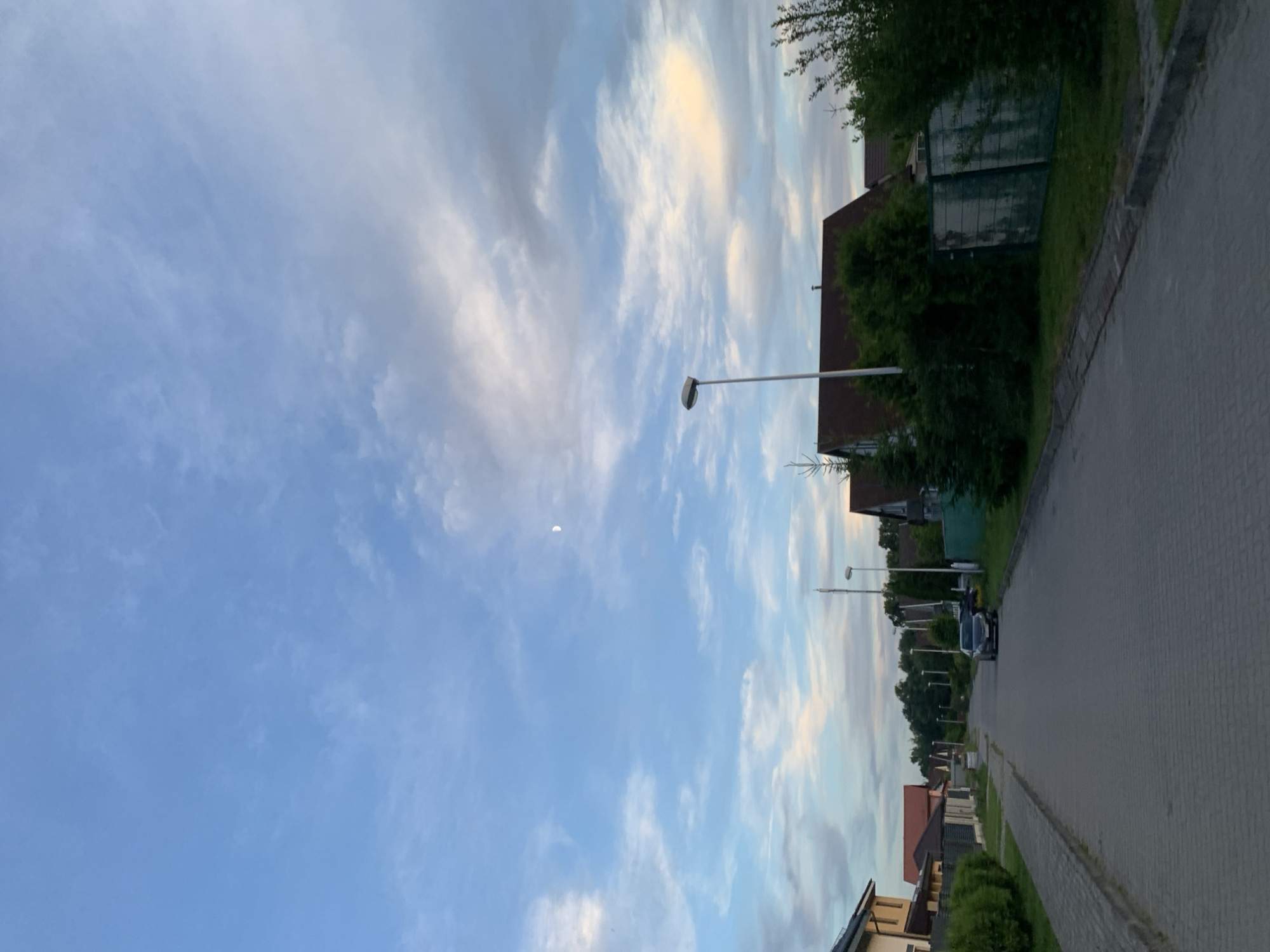 The image depicts a suburban street in Gvardeysk, Russia, under a twilight sky. The street is paved with interlocking gray stones, with a concrete curb and a paved sidewalk on the left. On the right side of the street, several houses with reddish-brown roofs are visible, interspersed with lush green trees and bushes. One house in the foreground has a light-colored facade. Further down the street, more houses and trees line the road, and several streetlights are present. A dark-colored car is parked on the right side of the road. The sky is a blend of pale blue and scattered white and gray clouds, with a faint crescent moon visible high up. The overall atmosphere suggests a peaceful evening in a residential area.