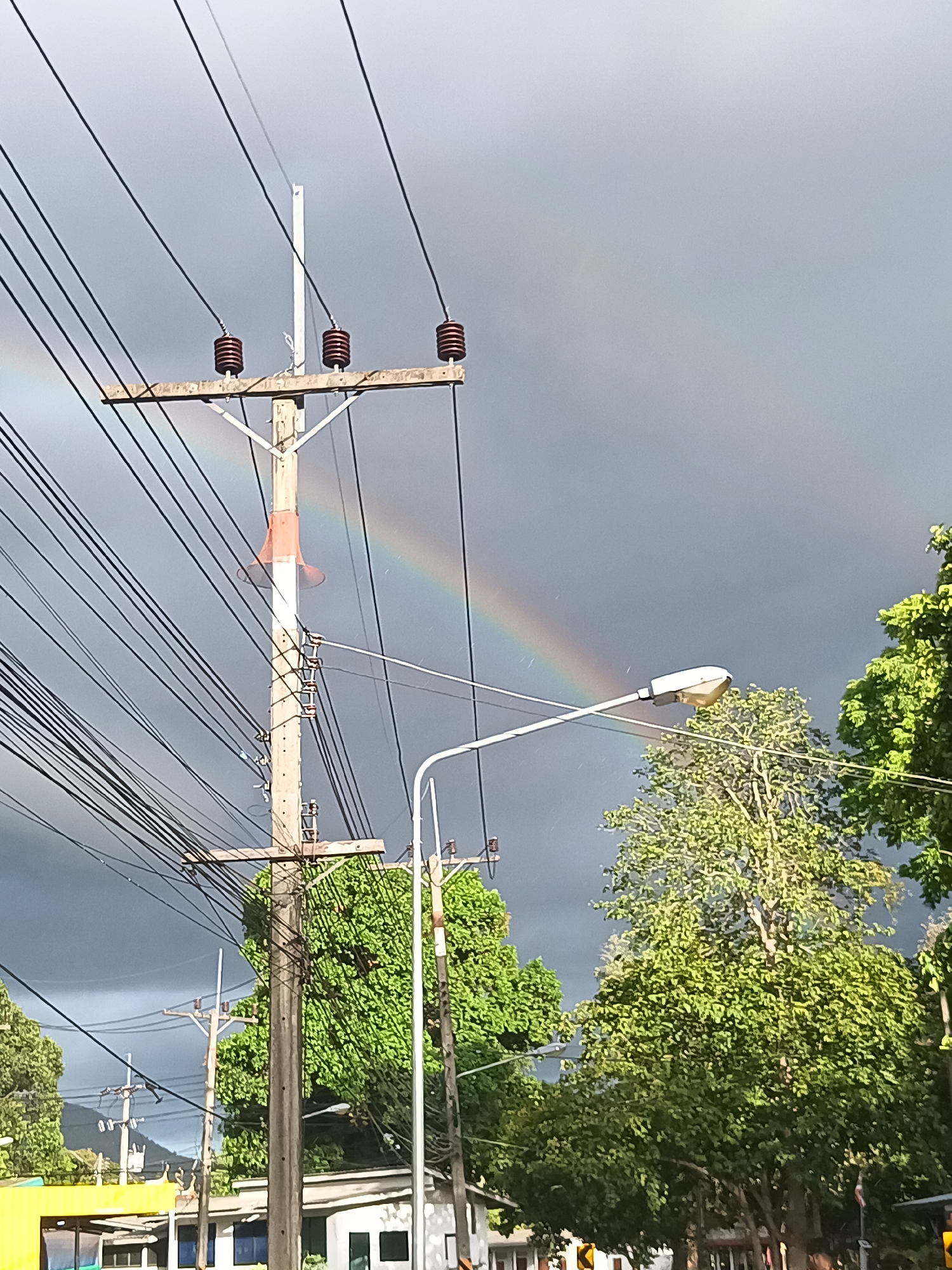 The photograph depicts a section of Moo 4, Thailand, under a predominantly overcast sky punctuated by a faint rainbow arc. A wooden utility pole, equipped with numerous power lines and insulators, stands prominently in the foreground. An attached red megaphone faces upwards. A street lamp illuminates the surrounding area. Trees with lush green foliage are interspersed throughout the scene. In the distance, other utility poles and buildings are visible.