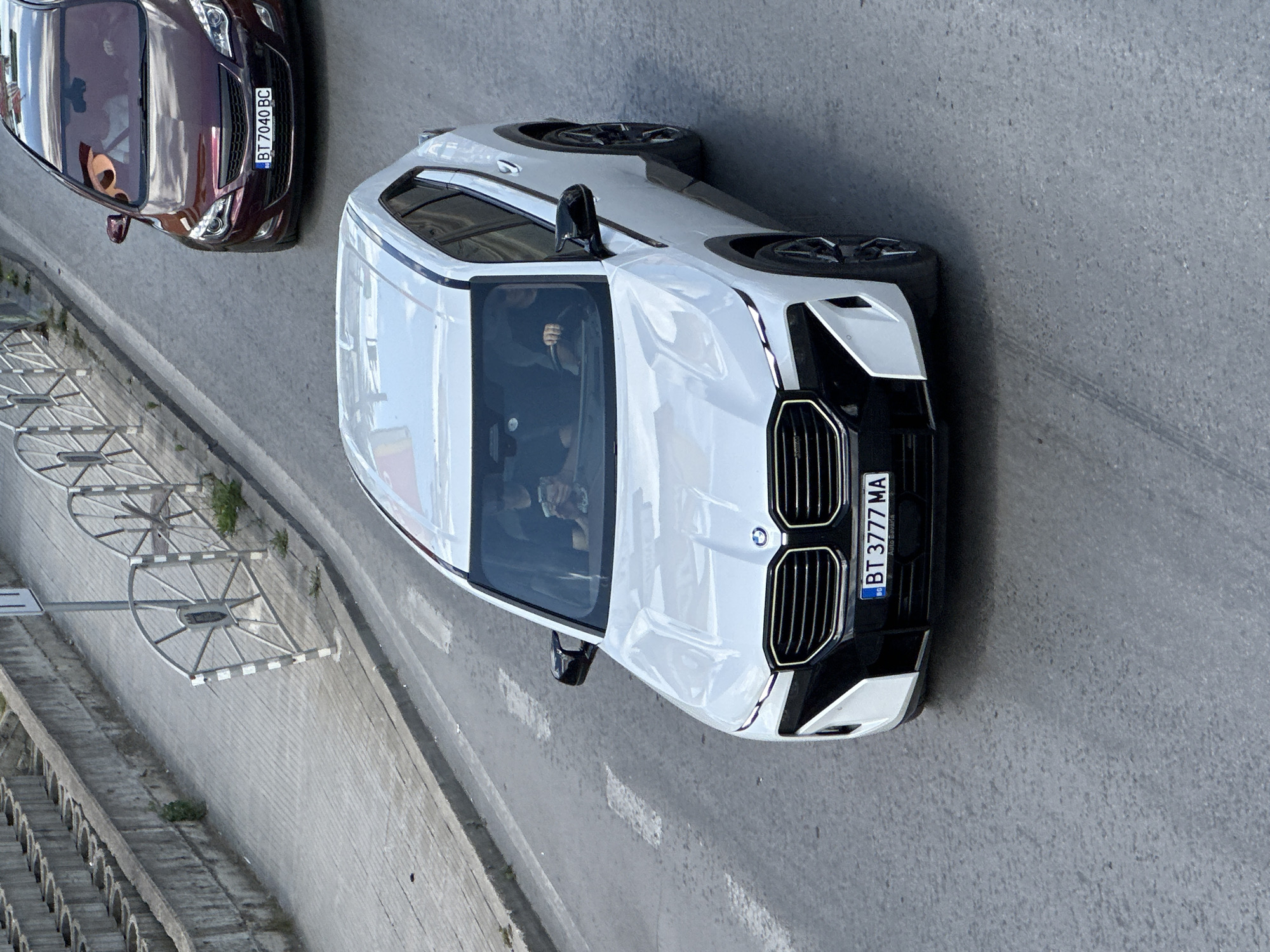 The image shows a white BMW X5 M parked on a street in Veliko Tarnovo, Bulgaria. The car is facing forward, and its license plate reads "BT 3777 MA". A dark red car is parked behind the BMW, with its license plate reading "BT 7040 BC". The street appears to be a multi-lane road, and there is a sidewalk with a metal fence to the left of the cars. In the background, there are buildings and trees, suggesting an urban environment. The weather appears to be overcast. The image is taken from a high angle, looking down on the cars.