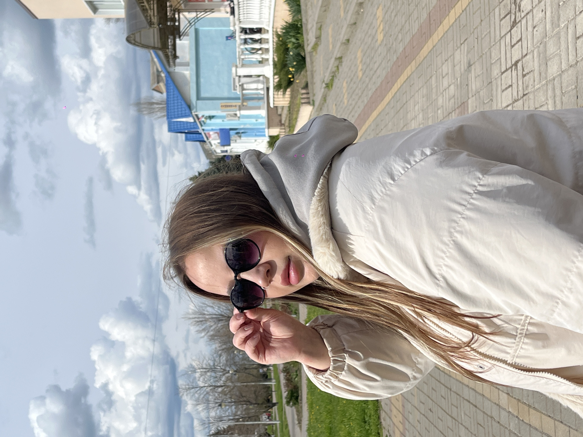 A young woman with long brown hair and wearing sunglasses stands on a sidewalk in Novopokrovskaya, Russia. She is wearing a light-colored hooded jacket. The sidewalk is made of rectangular paving stones. In the background are trees, grass, and buildings, including a light blue structure. The sky is cloudy.