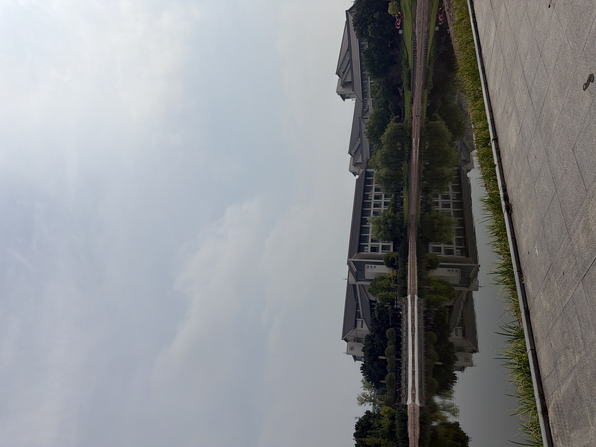 The image shows a wide shot of a lake with buildings and trees in the background. The sky is overcast. In the foreground, there is a paved walkway with some grass growing along its edge. The lake reflects the buildings and trees. The buildings appear to be modern in style with dark roofs. There are no people visible in the image. The scene suggests a park or a campus setting. The provided information states the location is in the city of Jiangjin, China.