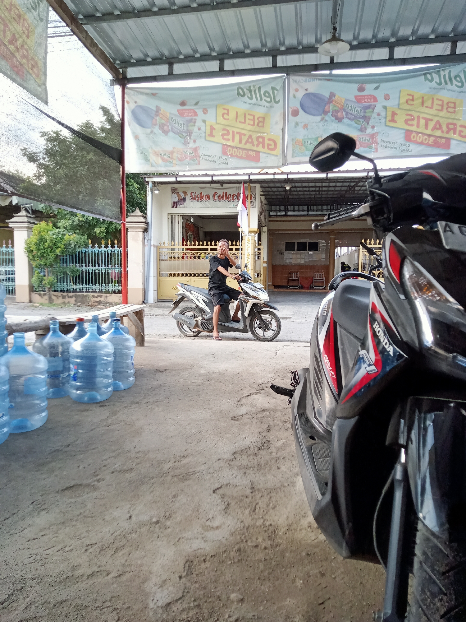 A man is seated on a white and black motorcycle in front of a storefront in Simo, Indonesia. The man, wearing a black t-shirt and shorts, is looking towards the camera and holding his right hand to his ear, possibly on a phone. The motorcycle is parked on a dirt and gravel surface. In the foreground, several large, blue plastic water jugs are arranged in a row. Behind the man and the motorcycle, the storefront has a red and white Indonesian flag displayed prominently, along with banners advertising what appear to be food items. The building itself has a corrugated metal roof extending over the entrance area. The overall environment suggests a roadside business or market stall.