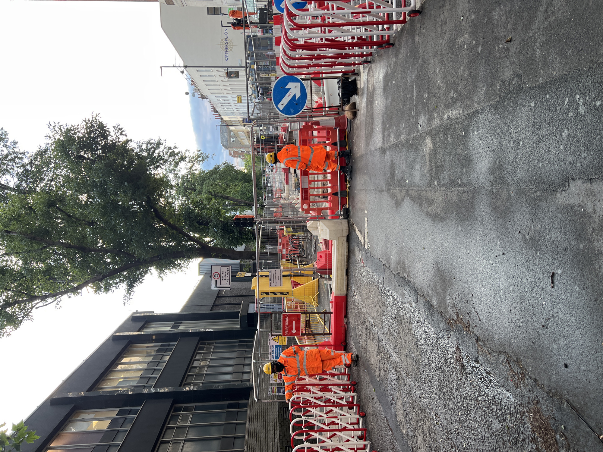 The image shows two construction workers in orange high-visibility suits and hard hats standing on a wet asphalt road. The road is partially blocked by red and white portable barriers and orange plastic safety fences. One worker is on the left side of the frame, leaning on a barrier, while the other is on the right, facing away from the camera. There are traffic lights and signs indicating "PEDESTRIANS" with an arrow pointing left. In the background, there is a modern building with dark tinted windows on the left, and a row of white buildings with shops and balconies on the right, including a sign for "WETHERSPOON". Trees line the street, and the sky is overcast. The scene appears to be a street in a city undergoing construction.