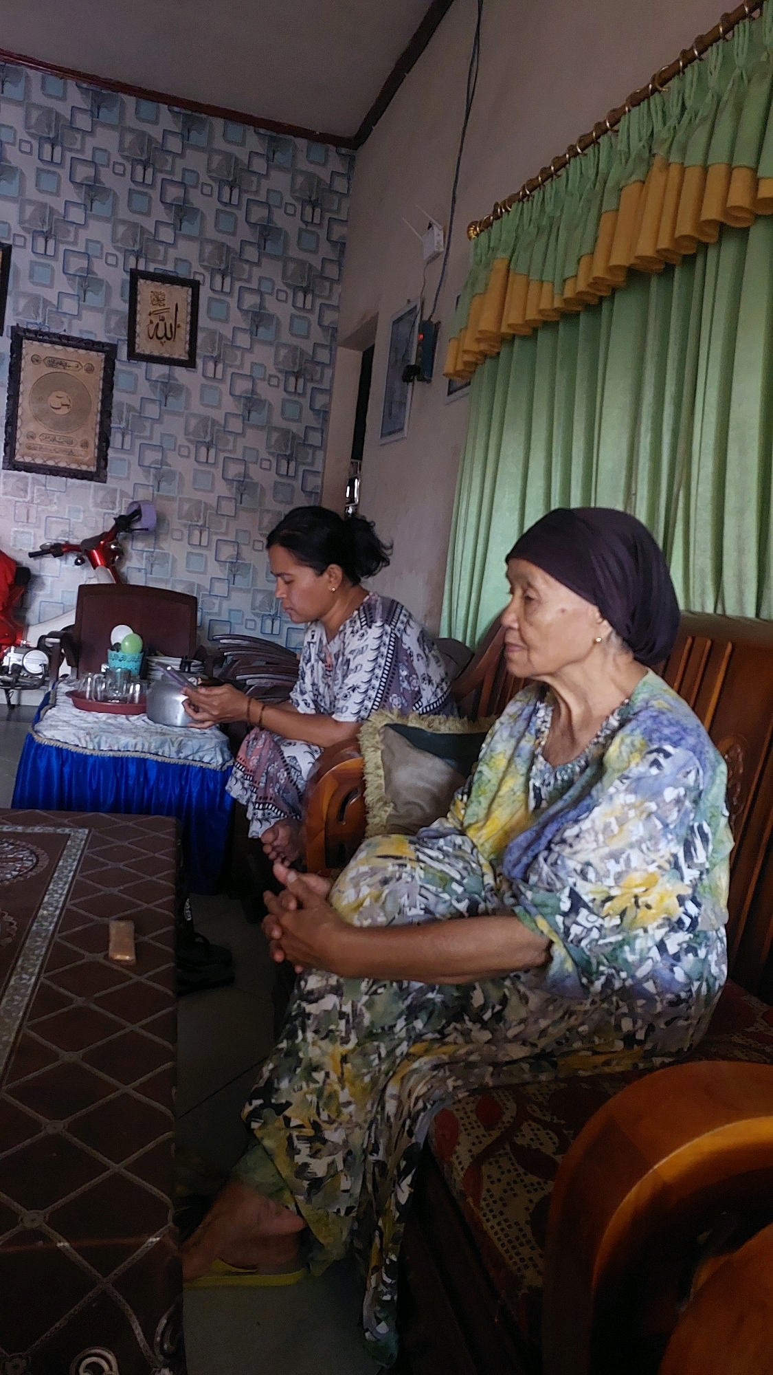 In a room with patterned wallpaper, two Indonesian women are seated. The woman on the left, wearing a patterned outfit, looks down at her phone. The woman on the right, wearing a headscarf and a colorful patterned dress, is looking to her left. A wooden bench is behind them, and a coffee table with a checkered tablecloth and a tray of items is in front of the woman on the left. The room appears to be in a residential setting in Sukowono, Indonesia, suggested by the style of furnishings and decor.