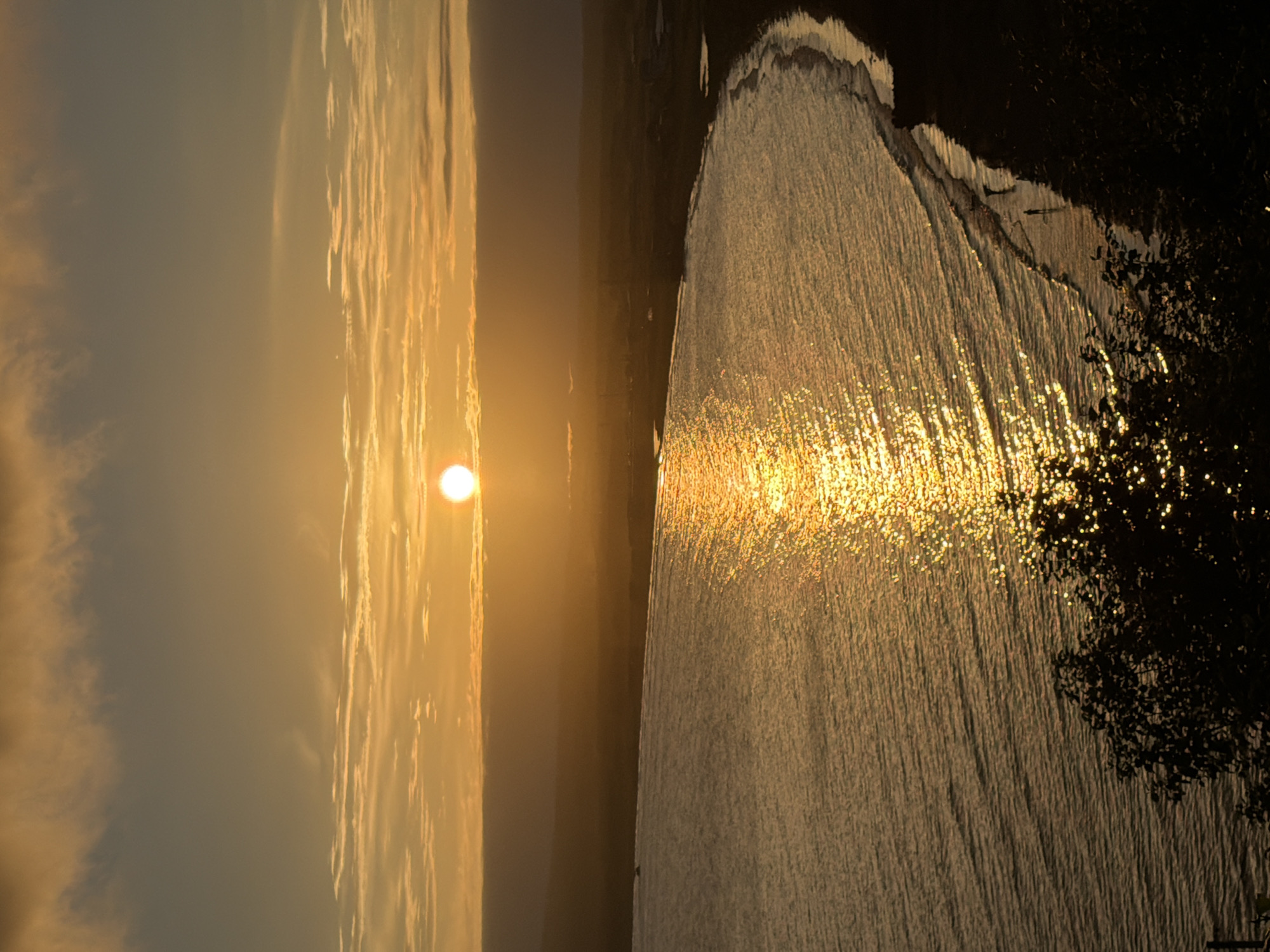 An aerial, medium-shot captures a sunset over the coast of Yuzhno-Kurilsk, Russia. The sun is a bright white orb near the center of the frame, partially obscured by thin, horizontal clouds tinged with orange and yellow. The sun's reflection creates a shimmering, golden path across the gently rippling water of the bay. The coastline curves from the right foreground, becoming a dark silhouette against the water. Small figures, appearing to be people, are visible walking along the wet sand at the water's edge. In the foreground, the dark silhouette of trees with glistening leaves from the sunset light adds depth to the scene. The overall impression is one of peaceful twilight.