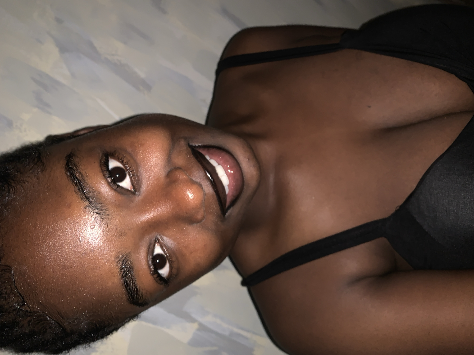A close-up, high-angle shot shows a Black woman with her head tilted to her left, looking directly at the camera. She has dark skin, dark eyes, and a wide smile, showing her teeth. Her lips are dark. Her eyebrows are dark and defined, and her eyelashes are long. Her hair is styled back, with some gelled edges visible on her forehead. She is wearing a black bra. The background is a textured wall with irregular blue and white brushstrokes. The lighting is warm and seems to be coming from the front, highlighting the sheen on her skin. The overall impression is of a casual, posed portrait.