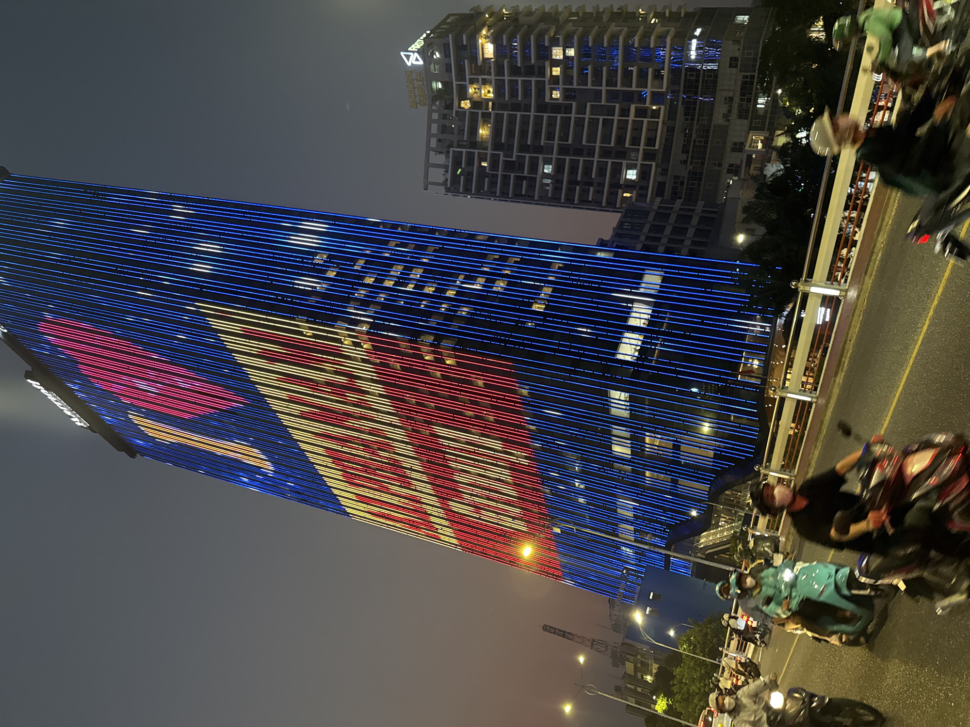 This nighttime shot captures the city of Đà Nẵng, Vietnam, with a focus on illuminated buildings and bustling street activity. A tall building is prominently featured with horizontal stripes of red, white, and blue lights. Another modern building with a unique geometric design is visible behind it. The foreground shows a busy street or bridge, with numerous scooters and motorbikes moving along the road. The environment is characterized by a dark sky and the glow of city lights. People are present, primarily as scooter drivers or passengers. The overall scene conveys the energetic urban atmosphere of Đà Nẵng at night.