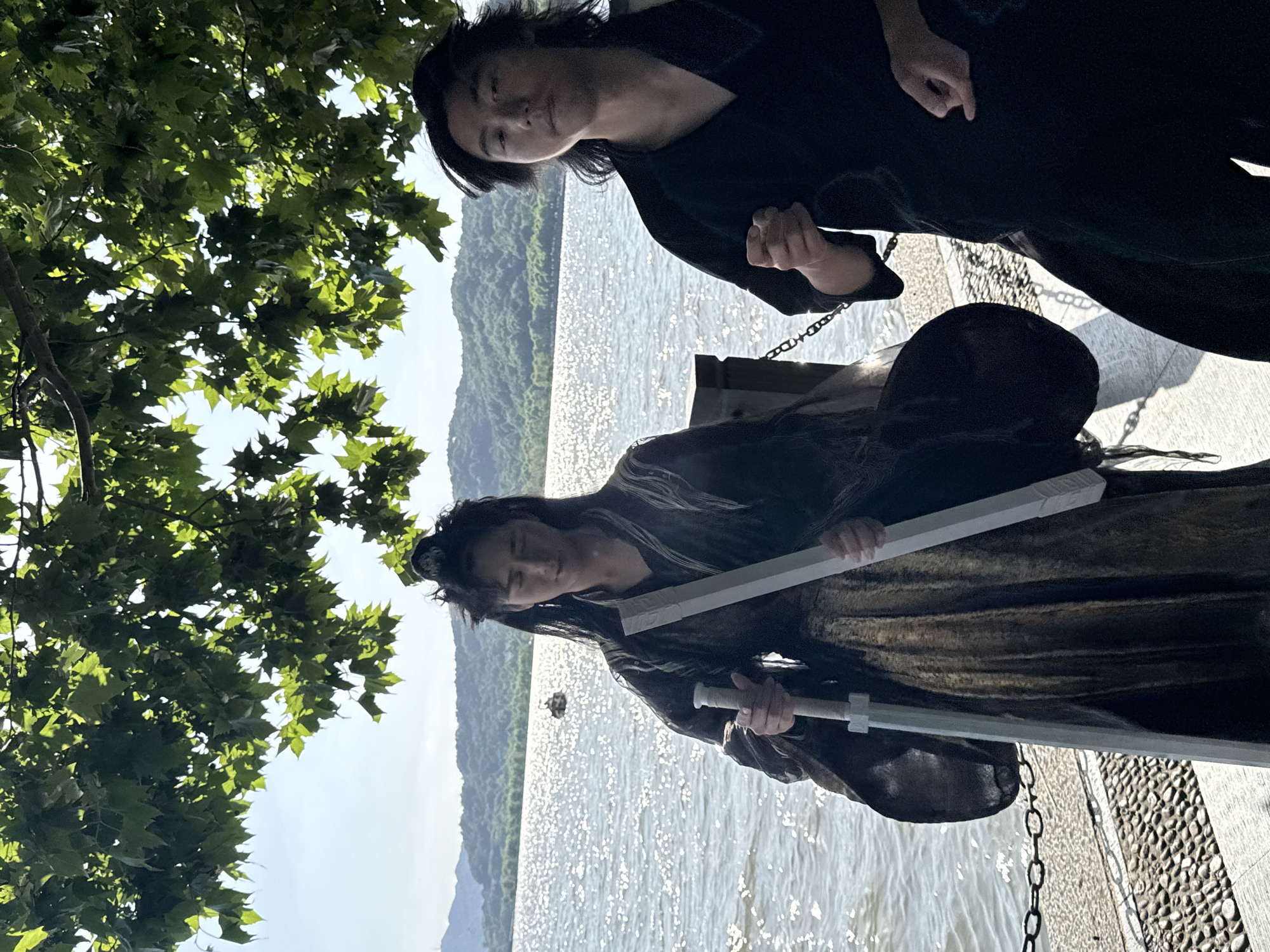 Two individuals are visible in this outdoor setting, likely within the Shangcheng District of China, given the provided context. The scene is captured on what appears to be a sunny day near a body of water, possibly a lake or river, with visible reflections on the surface.
One individual is positioned higher in the frame, wearing dark clothing. Their pose is casual, and they appear to be looking toward the camera. The second individual, situated lower in the frame, wears a dark outfit and holds a white, cylindrical object, possibly a prop such as a sword. The person seems to be in mid-action, perhaps posing or acting for a scene.
The background includes lush greenery, suggesting a park or natural area along the waterfront. A stone walkway or dock is visible, with metal chains along the edge, and tree leaves are to the left.