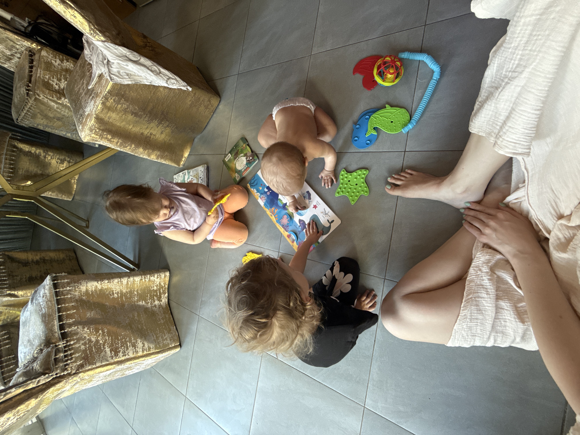 The scene is an indoor setting, likely a home, featuring a gray tiled floor. Three young children are the primary subjects, engaged with a book and toys. One child, wearing a diaper, is leaning over a book. Another child, wearing a lavender top and orange pants, is sitting to the side of the book. The third child, wearing black pants with white floral patterns, is also focused on the book. A woman, presumably the mother or caretaker, is seated on the floor with legs crossed wearing a white dress. Near the woman and the child in the diaper are colorful infant toys. The objects in the environment include gold textured seating, which gives context to the type of room. No context is given to determine location of this scene within the city of Чёрное, Russia.