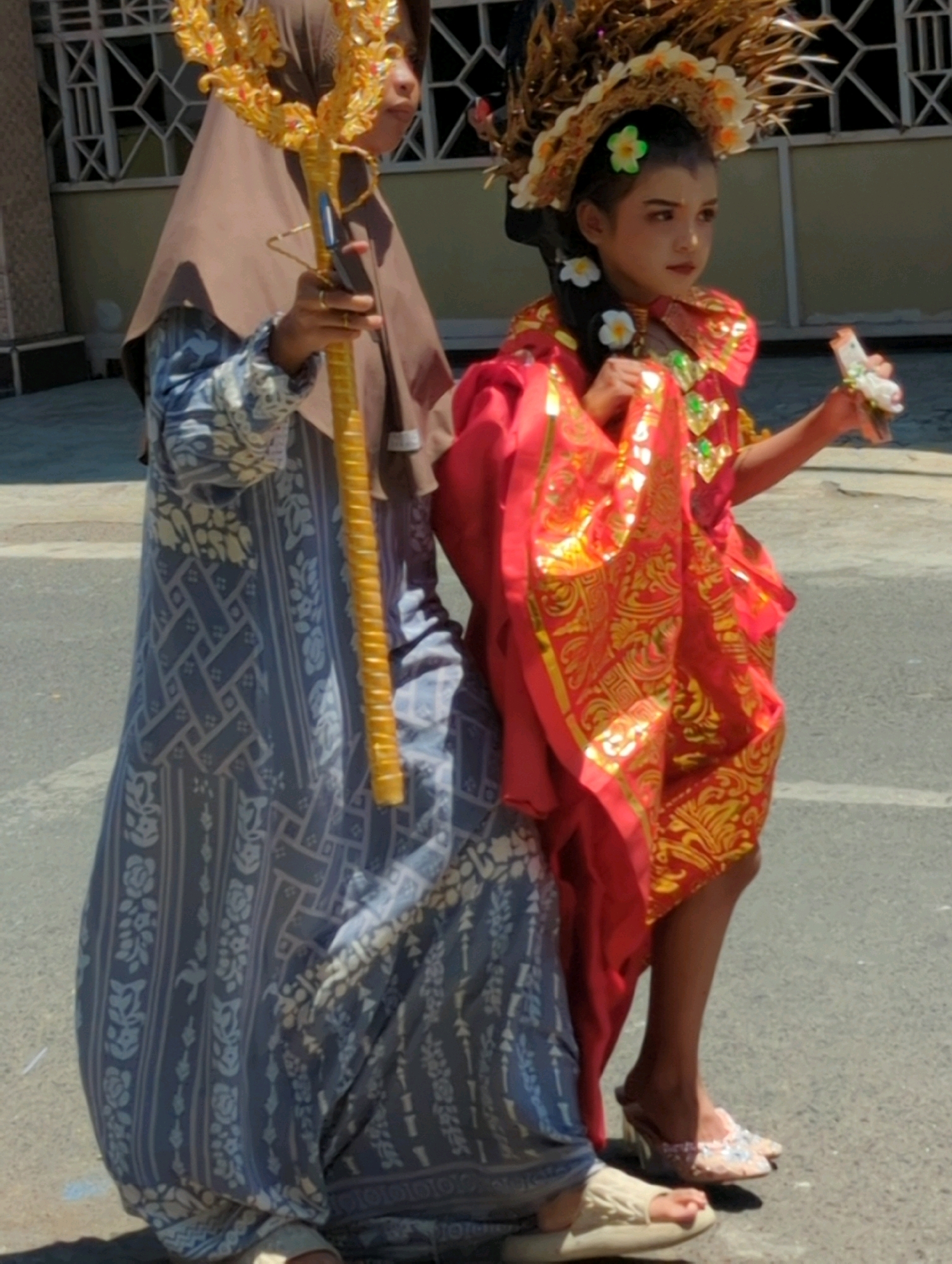 Two individuals are shown in an outdoor setting, possibly a street in Pasuruan, Indonesia, as suggested by the architecture visible in the background which resembles typical Indonesian urban construction. The person on the left, partially visible, wears a long, blue patterned garment and a brown hijab. They are holding a tall, ornate golden staff. To the right, a young person is adorned in a vibrant red and gold costume, featuring intricate patterns and decorative elements. They wear a headdress adorned with flowers and a necklace. This individual is holding a small, white object in their right hand, possibly a flower or a token. Both individuals appear to be walking on a paved surface, with a building with a barred window structure visible behind them. The lighting indicates it is daytime.