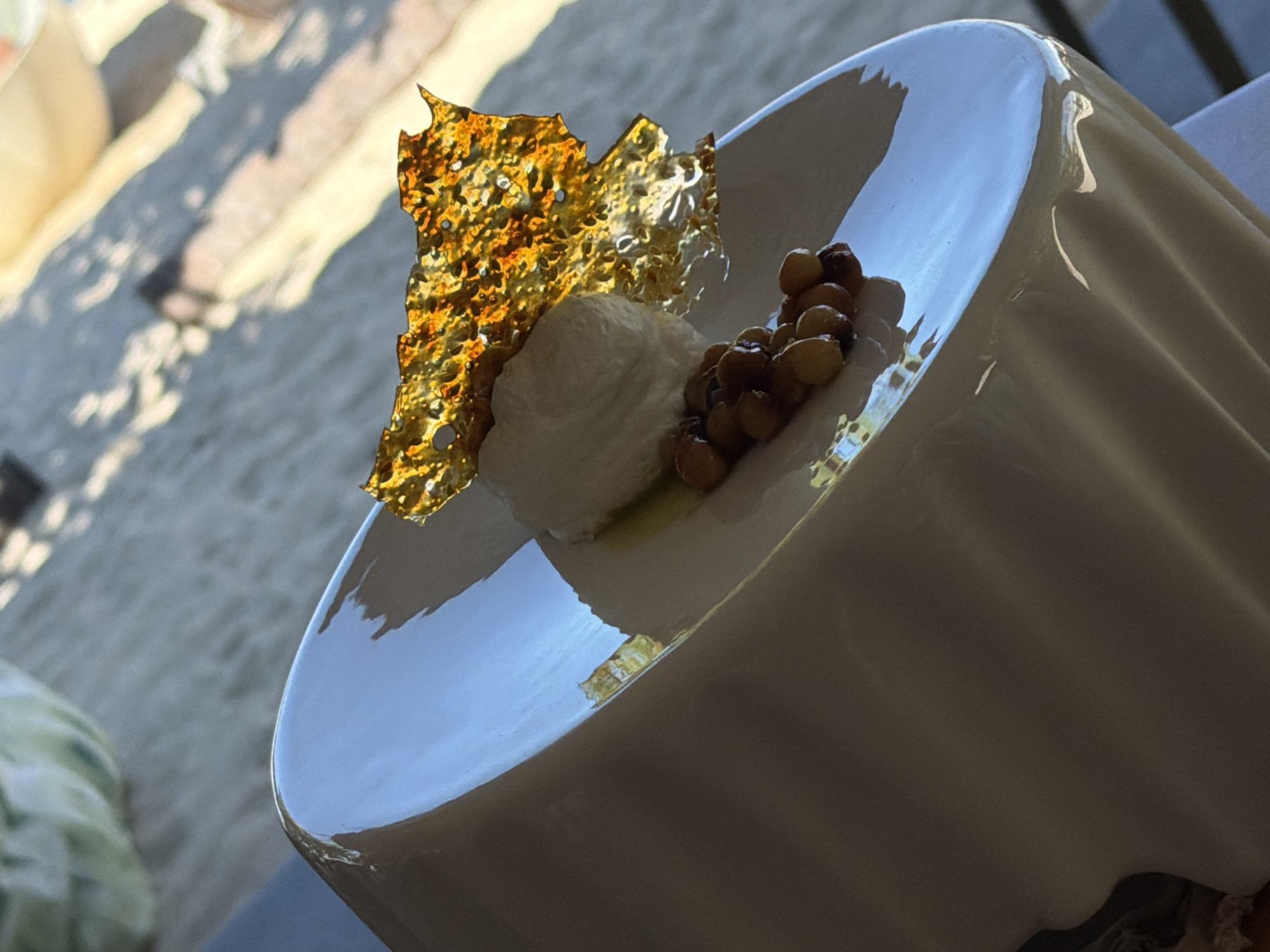This image features a close-up of a dessert served in a white, curved bowl. The dessert appears to be a scoop of white ice cream or sorbet, topped with a shard of golden, brittle candy or tuile. A small cluster of roasted nuts, possibly hazelnuts or almonds, is placed next to the ice cream. The bowl itself has a glossy finish and is set on a surface that reflects the light. The background is out of focus, showing a sandy or gravelly ground with dappled sunlight, suggesting an outdoor setting. The lighting indicates it is daytime, possibly late morning or afternoon, with bright sunshine creating shadows. The overall presentation is elegant and sophisticated, implying a fine dining experience. There is no visible text. The scene is likely a restaurant or cafe in Baan Thong Krut, Thailand, given the tropical-like lighting and texture of the ground.