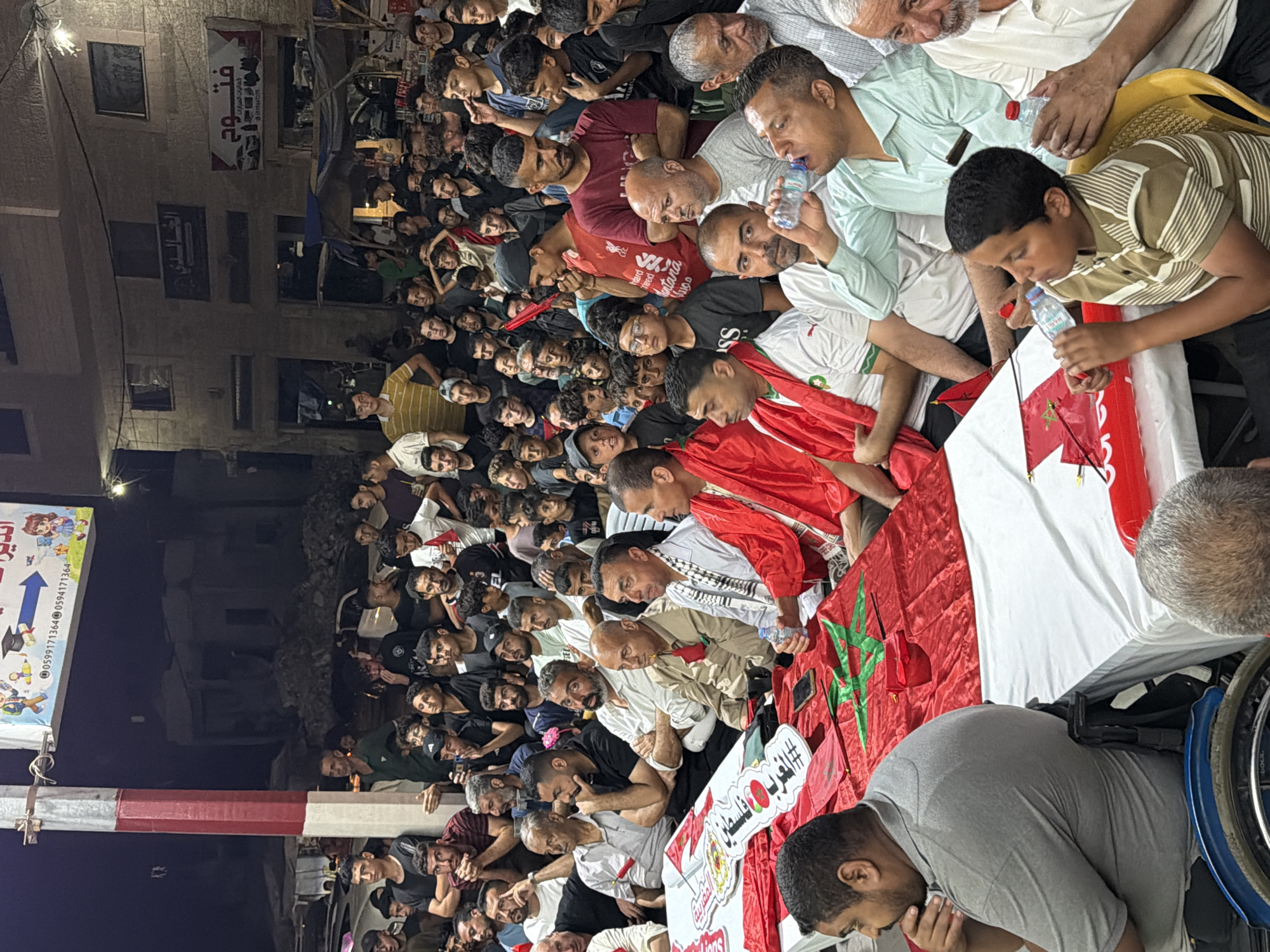 A gathering of men and boys, many wearing red and green clothing, are seated around tables covered with red cloths and Moroccan flags. Some individuals are holding small Moroccan flags on sticks, and a larger Moroccan flag is draped over one of the tables. The individuals appear to be watching or participating in an event. Behind the gathering, a building with multiple windows is visible, and a sign with Arabic script hangs on the wall. The scene suggests an outdoor gathering, possibly a celebration or a public event, in Gaza City, Palestinian Territories.