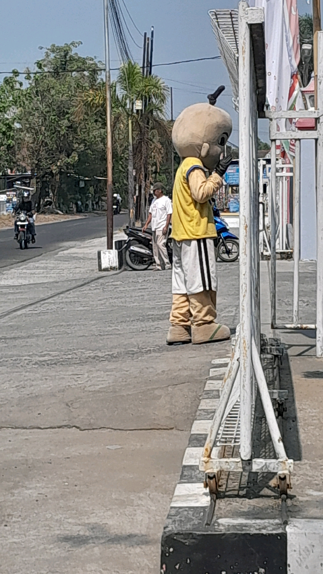 The image depicts a street scene in Tanjung, Indonesia, under a clear sky. In the foreground, a person in a mascot costume, designed to resemble a baby with a large head and a single dark tuft of hair on top, stands facing right, partially obscured by a white metal barrier. The costume is predominantly light brown for the head and legs, with a yellow long-sleeved shirt and white shorts with black stripes. The mascot is wearing large brown shoes and black gloves.
Behind the mascot, a man in a white t-shirt and beige pants stands next to a black motorcycle. Further in the background, a person rides a motorcycle on the road. Trees and utility poles line the street, and some buildings are visible in the distance. The road surface shows visible cracks. The overall impression is a bright, sunny day in an urban or semi-urban environment.