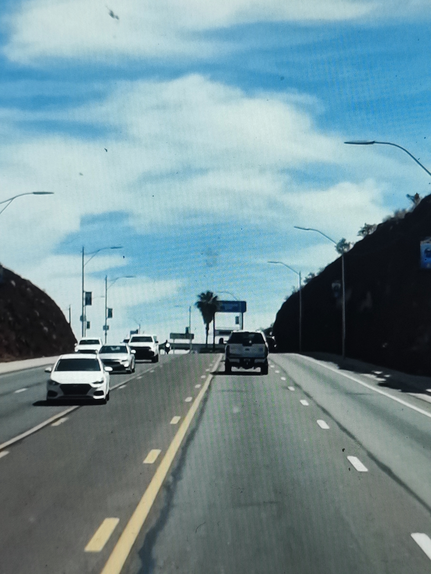 This image depicts a roadway with multiple vehicles traveling in the same direction. The scene appears to be a highway or a major road with multiple lanes. On the left side of the road, there is a rocky, mountainous terrain with vegetation. On the right side, the terrain is also hilly, with some trees visible. The sky is blue with scattered white clouds. Several streetlights are visible along the road. The vehicles in the image include sedans and what appears to be a truck or SUV. The perspective suggests the image was taken from a moving vehicle. There is no information within the image to suggest this location is in Yarmolyntsi, Ukraine.