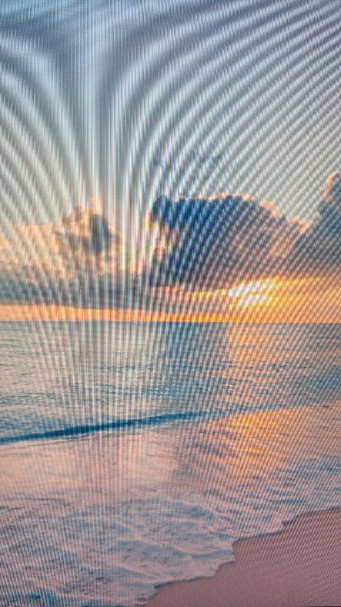 The image depicts a serene beach scene at sunset. The sky is painted with hues of orange, pink, and blue, with fluffy clouds illuminated by the setting sun. The ocean reflects these colors, creating a shimmering effect on the water's surface. Gentle waves lap at the sandy shore, which is visible in the foreground. There are no people or objects present in the image, creating a tranquil and naturalistic atmosphere.
There is no connection to the city of Kuznetsk, Russia.