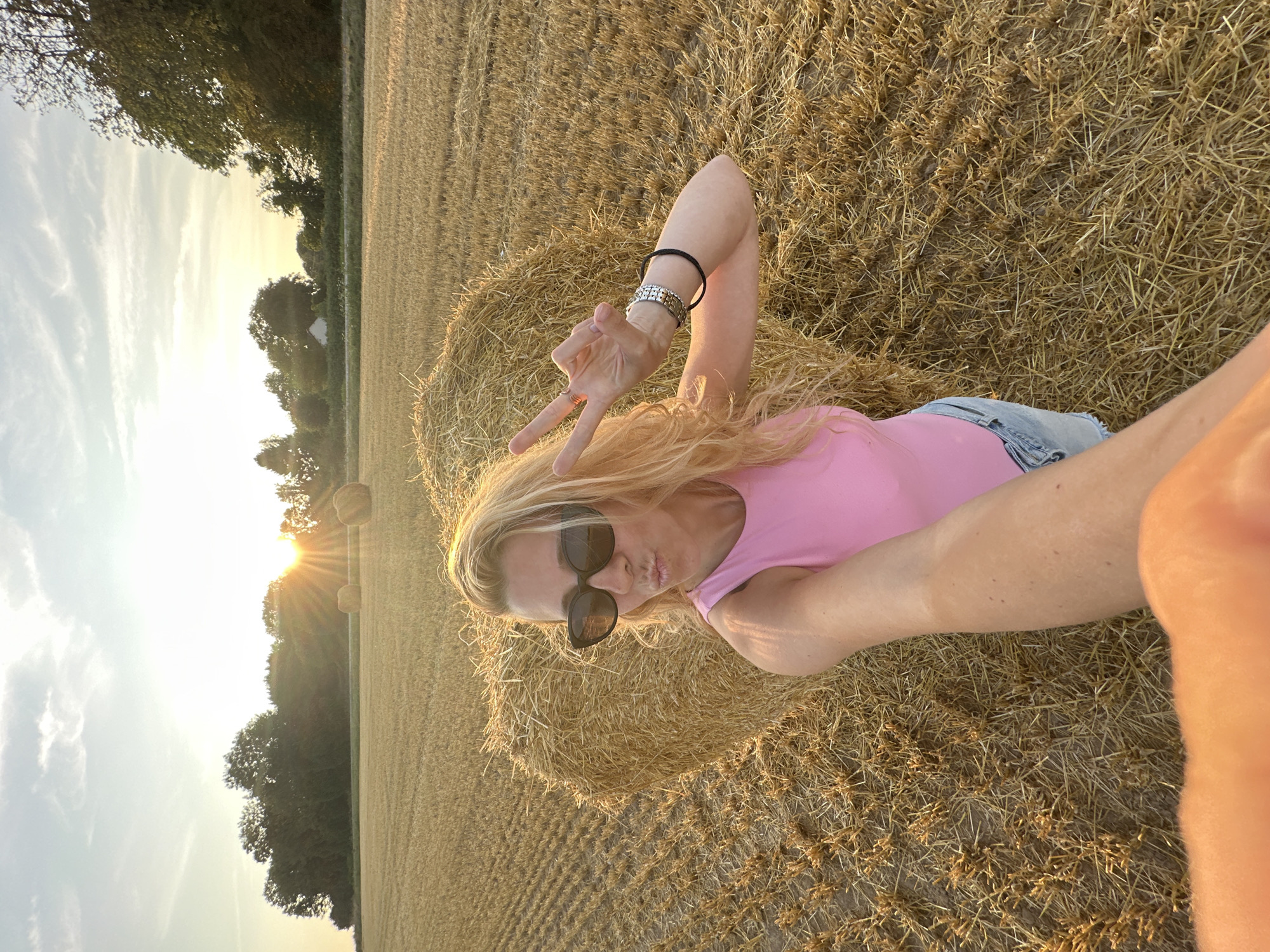 A blonde woman with long, wavy hair and sunglasses is taking a selfie in a harvested field. She is wearing a pink top and denim shorts. She is standing in front of a heart-shaped haystack, and a few other haystacks are visible in the background. The sun is setting behind a line of trees, casting a warm glow over the scene. The field is covered in stubble, indicating it has recently been harvested. The location is identified as Trouville-Alliquerville, France.
